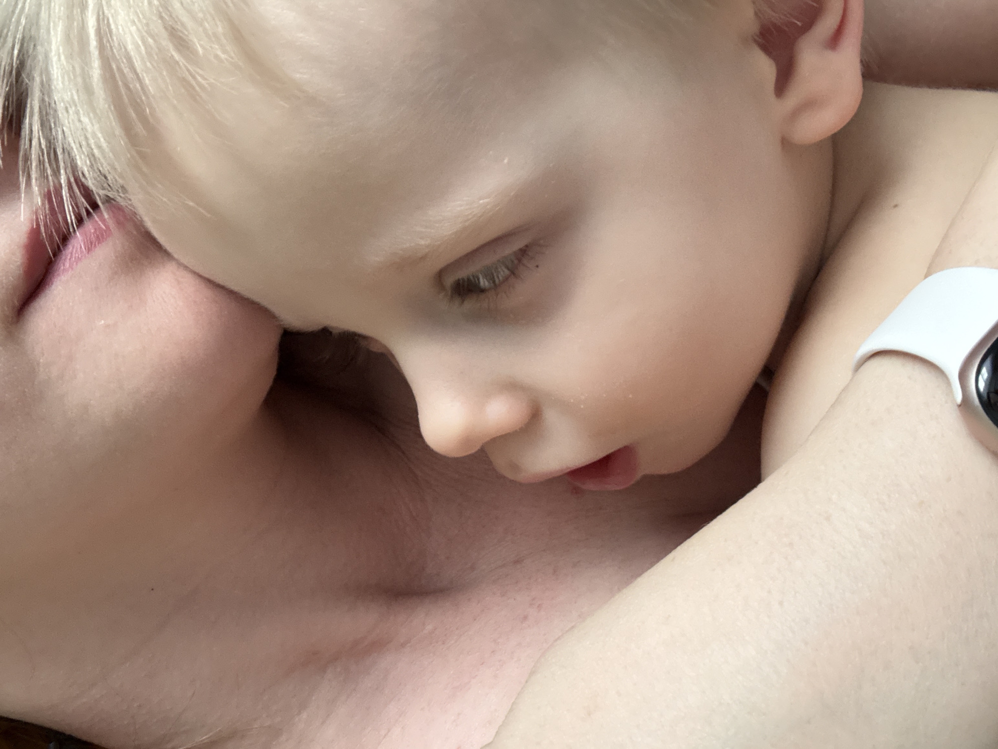 A close-up shot depicts a young child with blonde hair lying on an adult's chest. The child's face is turned slightly downwards, eyes looking down, and mouth slightly open. The adult's face is partially visible on the left side of the frame, showing a portion of their lips. The child's arm is draped across the adult, with a white wristwatch visible on their wrist. The setting of the image cannot be determined from the visual context. There is no indication of this image being taken in Chaikovsky, Russia.