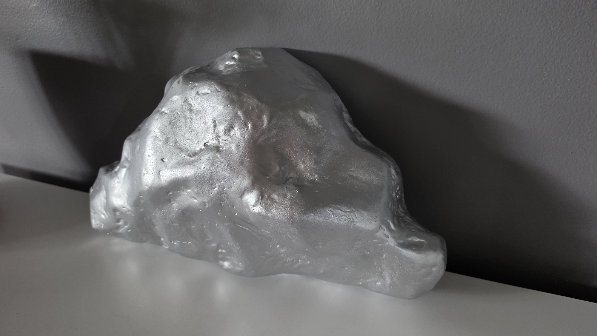 A metallic silver, amorphous object is placed on a white surface in front of a gray wall. The object has a rough, textured surface with visible undulations and bumps, giving it the appearance of a cooled metallic mass or a sculpted rock. Light reflects off the metallic surface, creating highlights and shadows that emphasize its three-dimensional form. A distinct shadow is cast on the gray wall behind the object, originating from a light source to the left of the frame. The background wall is textured and appears to be a solid color. The overall scene is lit, with the primary light source coming from the left, casting a prominent shadow. There is no indication of the city of Elgin, United States of America, in this image.