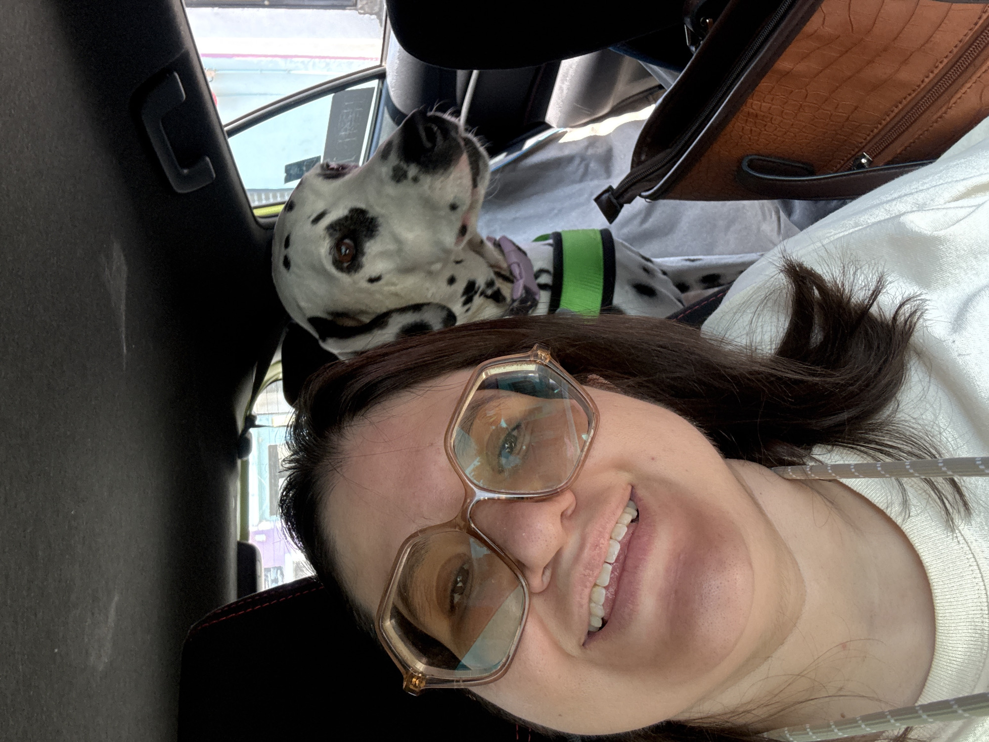 A woman takes a selfie from the passenger seat of a car, with her Dalmatian dog positioned beside her. The image is taken from a slightly low angle, focusing on the woman's smiling face in the foreground, and the dog in the midground.
The woman has dark hair and is wearing distinctive wide-rimmed, light-colored hexagonal glasses, through which her light brown eyes are visible. She is smiling broadly, showing her teeth, and appears to be wearing a white ribbed top, possibly a sweater, under a darker garment with red stitching visible on the shoulder. Her face is angled slightly, looking directly at the camera.
To the woman's left, partially obscured by her, is a black and white spotted Dalmatian. The dog is looking upwards and to the left, away from the camera, possibly out a window. It wears a green chest harness or vest over a purple collar. Its right eye is visible, and its nose is black.
The setting appears to be the interior of a car. Dark upholstery or interior panels are visible, including a black hook or holder on a pillar to the far left. In the background, behind the dog and slightly to the right, rests a brown, textured bag with an alligator-skin pattern and visible zippers. Outside the car window, hints of an urban environment with muted colors can be seen. The bright lighting suggests it is daytime. The scene evokes a feeling of companionship and travel in Xochitepec, Mexico.