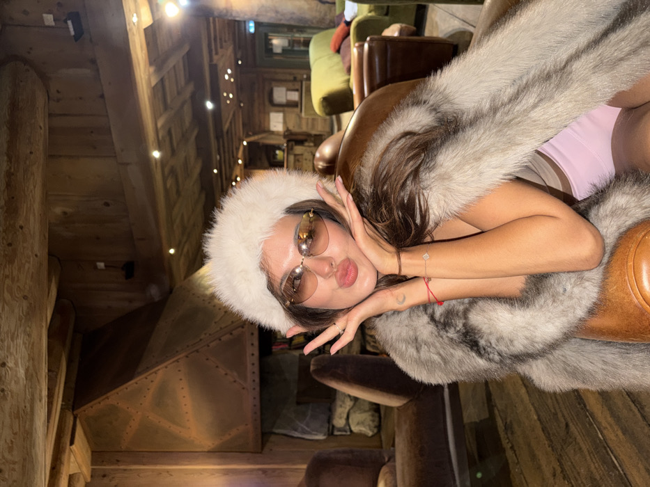 A woman poses indoors, clad in a fur hat and stole, lounging in a leather armchair. Her arms are raised to her face, hands framing it, while she puckers her lips. She wears rose-tinted sunglasses and bracelets on her wrists. The interior shows rustic wooden beams and walls, accented by string lights. A copper-colored structure sits in the background, hinting at a fireplace or ventilation system. Additional furniture is visible in the blurry background, suggesting a lounge or cabin setting.
