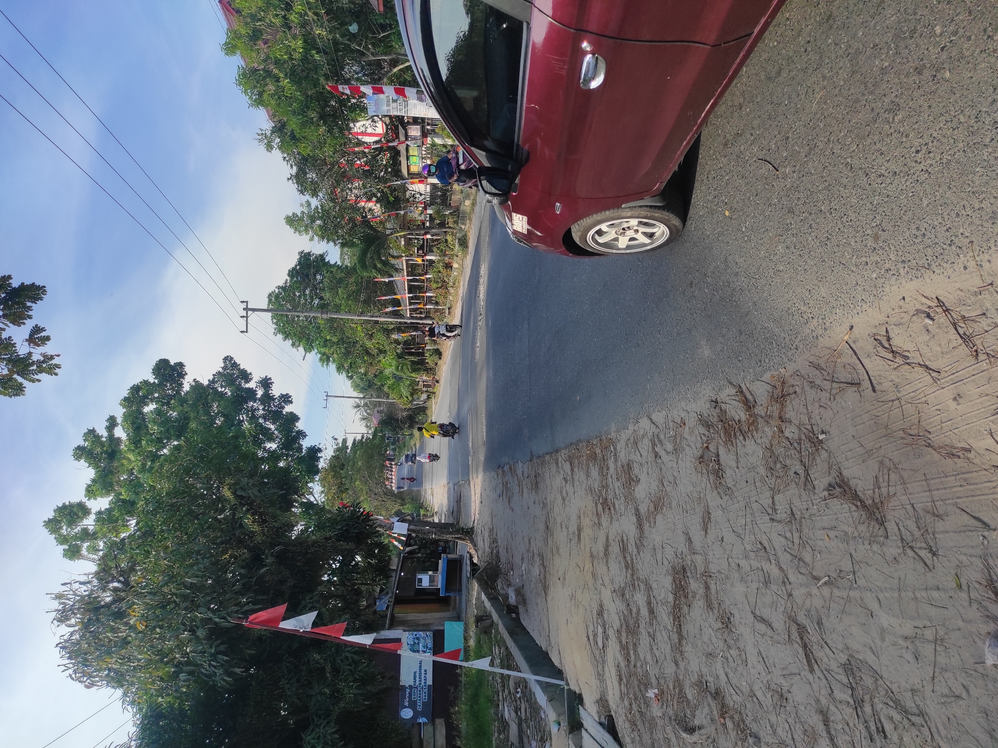 This is a medium-angle, eye-level, outdoor photograph taken in daylight on a paved road in the city of Muara Jawa Ulu, Indonesia. A maroon car is parked in the foreground, on the right side of the frame, facing away from the viewer. The car's front passenger-side wheel and a portion of the body are visible. The road continues into the distance, with several motorcycles and their riders traveling in the same direction as the car. Along the left side of the road, there is a sandy shoulder with some debris and vegetation. A small building with red and white flags is situated on the left, adjacent to the road. Trees with lush green foliage line both sides of the road. The sky above is blue with a few wispy clouds. Electrical wires are strung overhead, supported by utility poles.