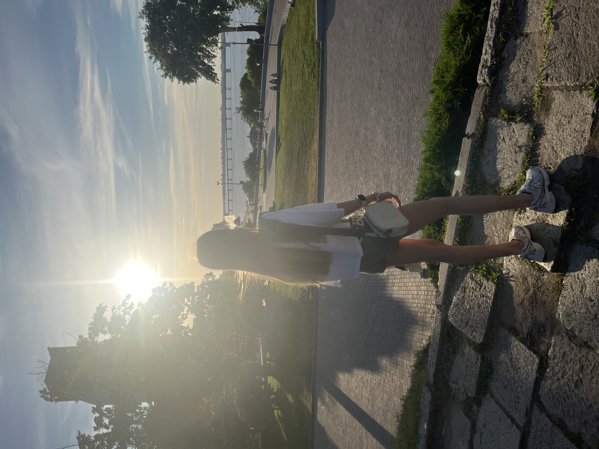 The image captures a young woman with long, blonde hair standing on a paved walkway next to a grassy area and large stone steps. She is wearing a white top, olive green shorts, and white athletic shoes. She carries a small white crossbody bag. In the background, a wide river is visible, with a bridge spanning across it. Trees line the riverbank and the walkway, casting long shadows. The sun is low in the sky, creating a strong lens flare and casting a warm, golden light over the scene, suggesting it is either sunrise or sunset. The sky is bright blue with wispy white clouds. The overall atmosphere appears serene and peaceful, hinting at a pleasant outdoor moment. The location is identified as Dnipro, Ukraine. There is no visible text in the image.