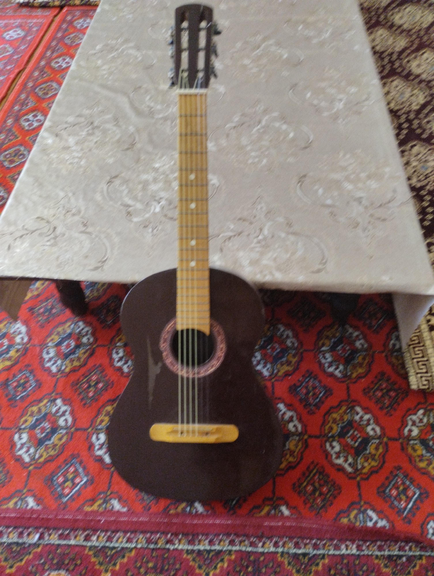 A brown acoustic guitar is placed upright on a patterned rug in front of a table covered with a textured cloth. The guitar has six strings and a light-colored fretboard with fret markers. The rug has a dominant red color with intricate geometric patterns in yellow, blue, and white. The table cloth is a light beige with an embossed swirling design. The scene appears to be indoors.