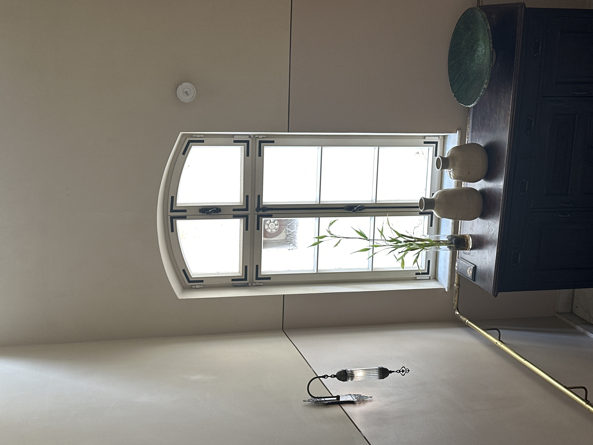 The image shows an interior scene with a focus on a window and adjacent elements. The window is multi-paneled, with rounded top sections on the left side. On the windowsill are two vases, a bowl, and a glass container holding green foliage. Next to the window is a dark, ornate cabinet. Below the window is a wall-mounted decorative light fixture. The ceiling is a light color, and a smoke detector is visible. There is no evidence to definitively place this scene in Tel-Aviv, Israel based solely on the provided image.