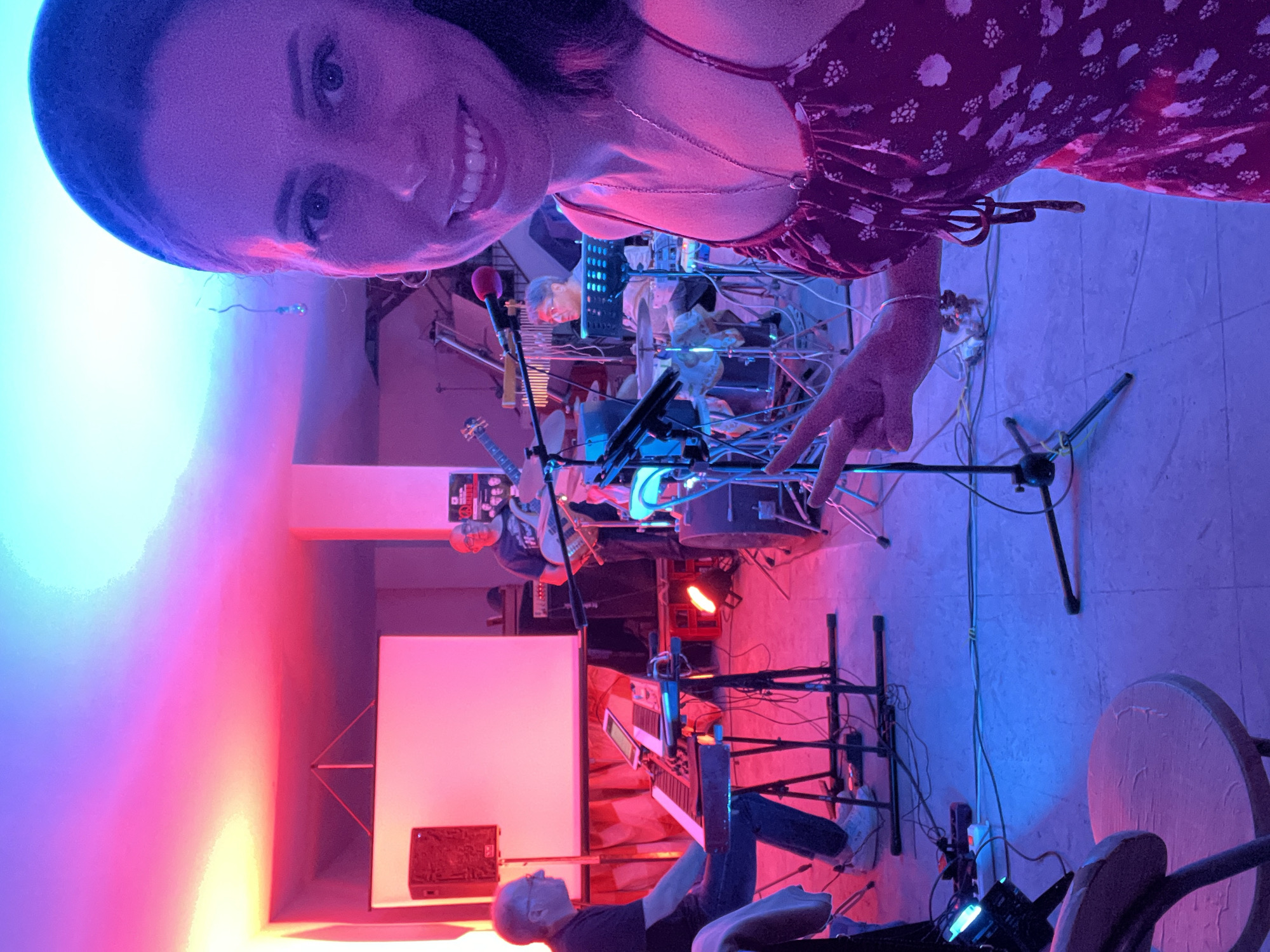 This image captures an indoor musical performance in Santeramo in Colle, Italy. The scene is illuminated by dominant red, blue, and purple colored lighting, which projects onto the light-colored walls and tiled floor.
In the immediate foreground, a woman is taking a selfie, smiling and looking directly at the camera. She is wearing a dark red dress with a white floral pattern and a silver chain necklace. Her right hand is raised, forming a V-sign (peace sign). Her face is primarily illuminated by blue and purple light. A round, light-colored wooden stool or table is partially visible in the lower-left corner.
Behind the woman, a five-piece band is actively performing. From left to right:
A male keyboardist, wearing glasses, a dark shirt, and light trousers, is seated and operating an electronic keyboard positioned on a stand. A tablet or piece of sheet music is visible on the keyboard. To his left, a red speaker cabinet stands on the floor. Behind him and extending to the right, a large white projection screen is suspended.
To the right of the projection screen, a male musician wearing glasses and a dark t-shirt (displaying a graphic) stands, holding a light-colored bass guitar. Behind him, partially obscured by equipment, a male saxophonist is playing a saxophone.
Further to the right and in the background, a male drummer, also wearing glasses, is seated and playing a drum kit.
The performance area is populated with multiple microphone stands, various audio cables running across the floor, and stage lighting units. A white power strip is visible on the floor near the foreground equipment. A dark poster or banner, featuring a peace sign graphic, is affixed to the wall behind the drummer. The ceiling above the performance space displays a gradient of blue, pink, and purple light.