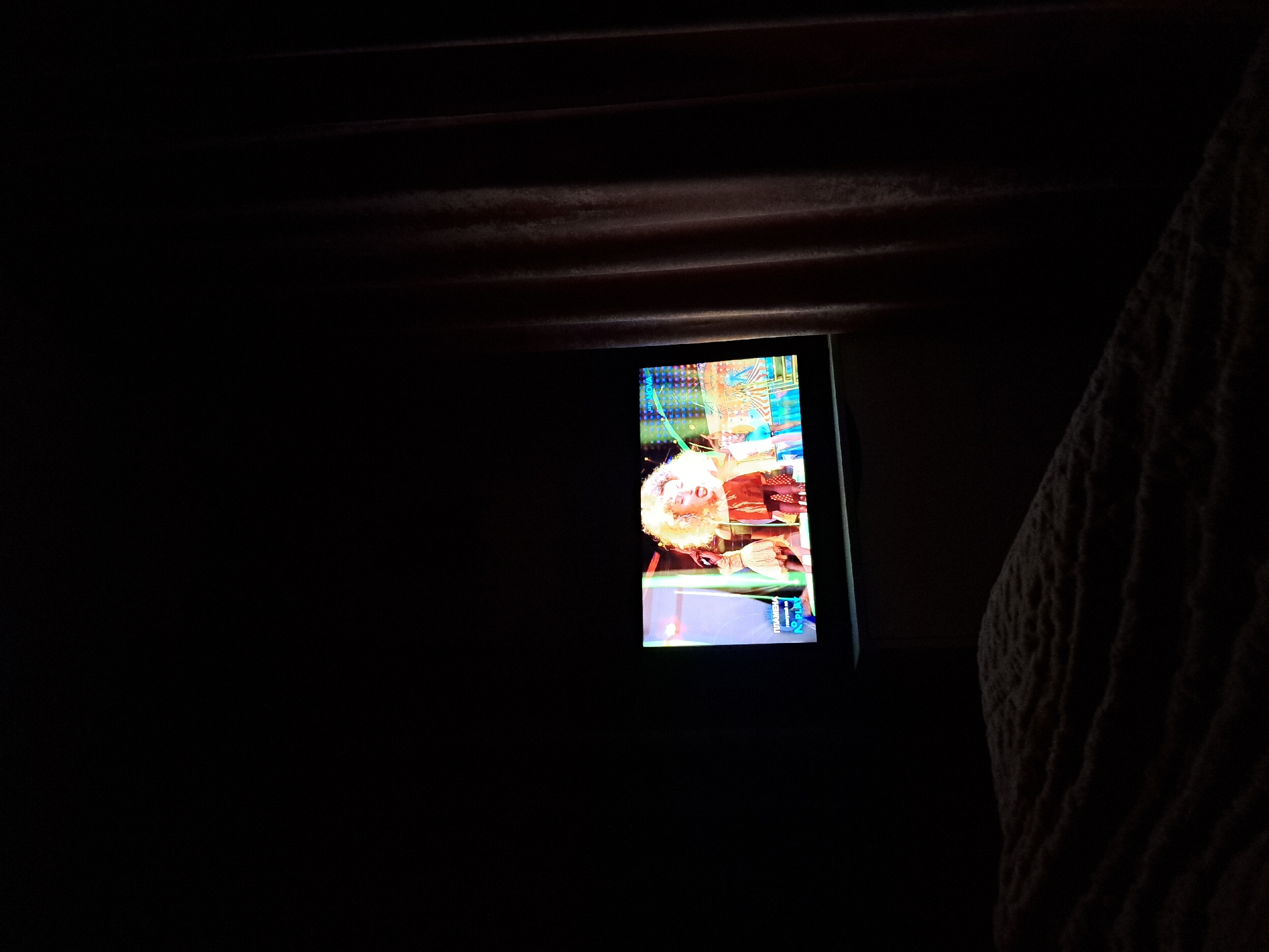 In a dimly lit room in Chepintsi, Bulgaria, a television screen is the primary source of light. The screen displays a person with blonde hair, a patterned outfit, and what appears to be a music video or performance. The room is otherwise dark, with dark curtains visible at the top and a textured, dark fabric on the right side.