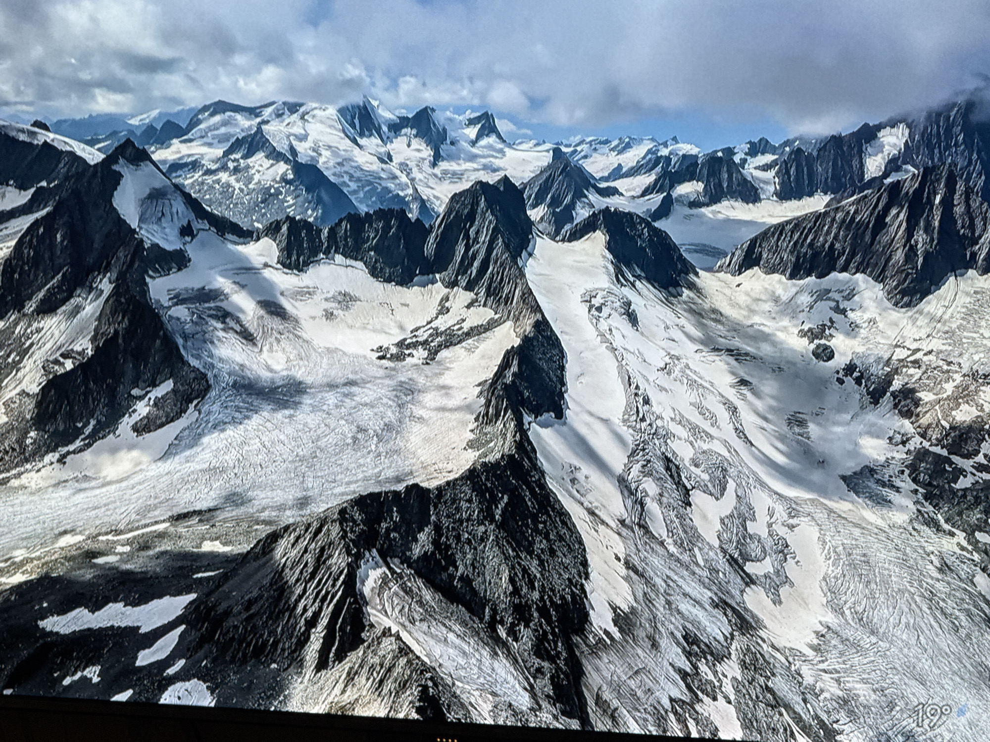 The image depicts a vast, high-altitude mountain landscape covered in snow and glaciers. Jagged, dark rock peaks rise sharply above extensive ice fields that show evidence of crevasses and moraines. The sky above is a mix of blue with scattered clouds. In the bottom right corner, a small digital display shows "19°" and a circular icon, possibly indicating temperature and weather. The scene is a natural, mountainous environment, not within the city of Saint Petersburg, Russia.