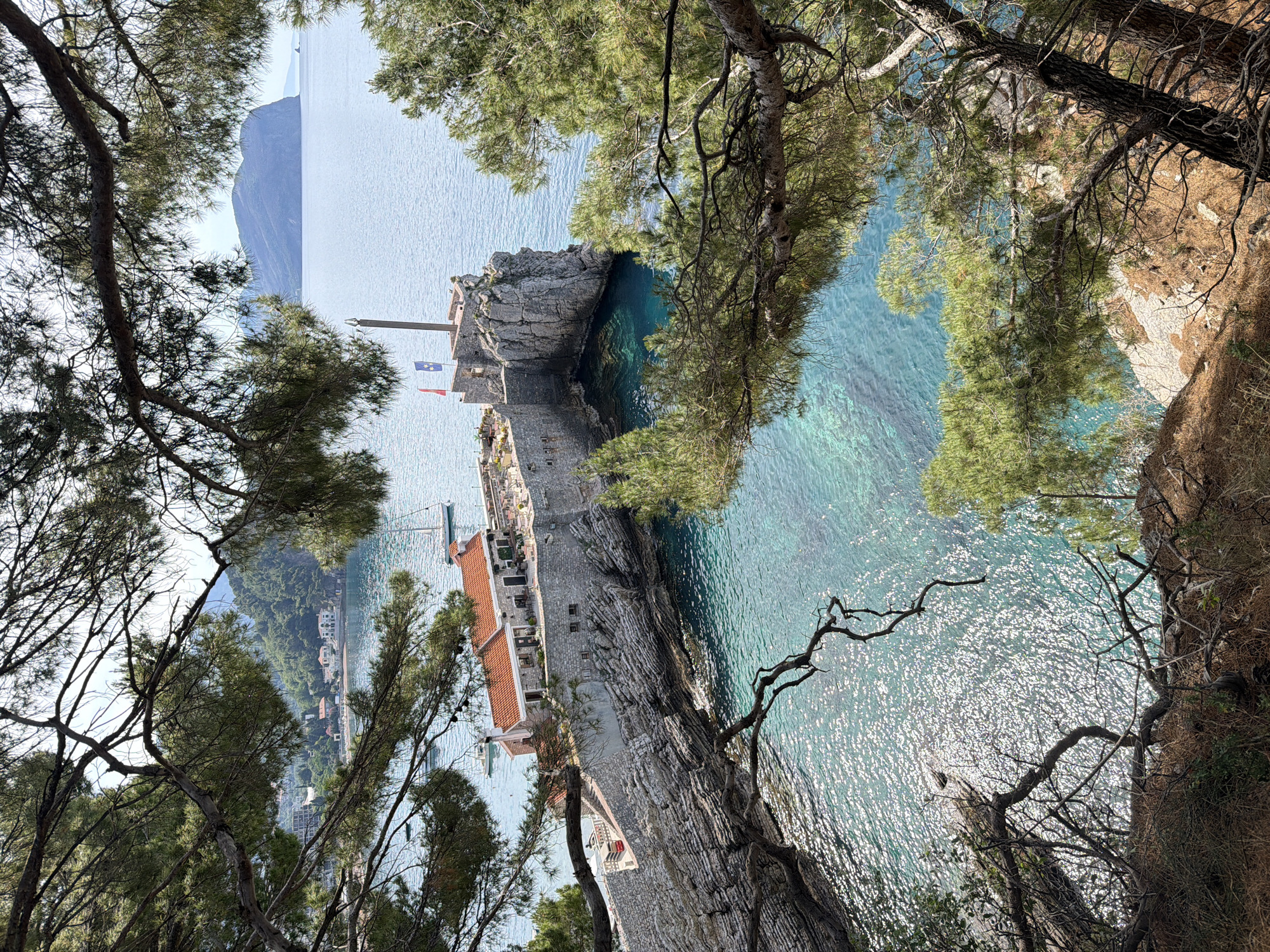 A high-angle shot shows a coastal view in Petrovac na Moru, Montenegro. In the distance, a large, hazy mountain emerges from the Adriatic Sea. Closer, the image features a stone pier with a few buildings with terracotta rooftops at its base. One structure displays two flags on a pole. The turquoise waters of the sea surround the pier, and sunlight glints off the surface. In the foreground, tree branches frame the view from above, adding depth and perspective. The surrounding landscape is a mix of rocky cliffs and dense foliage.