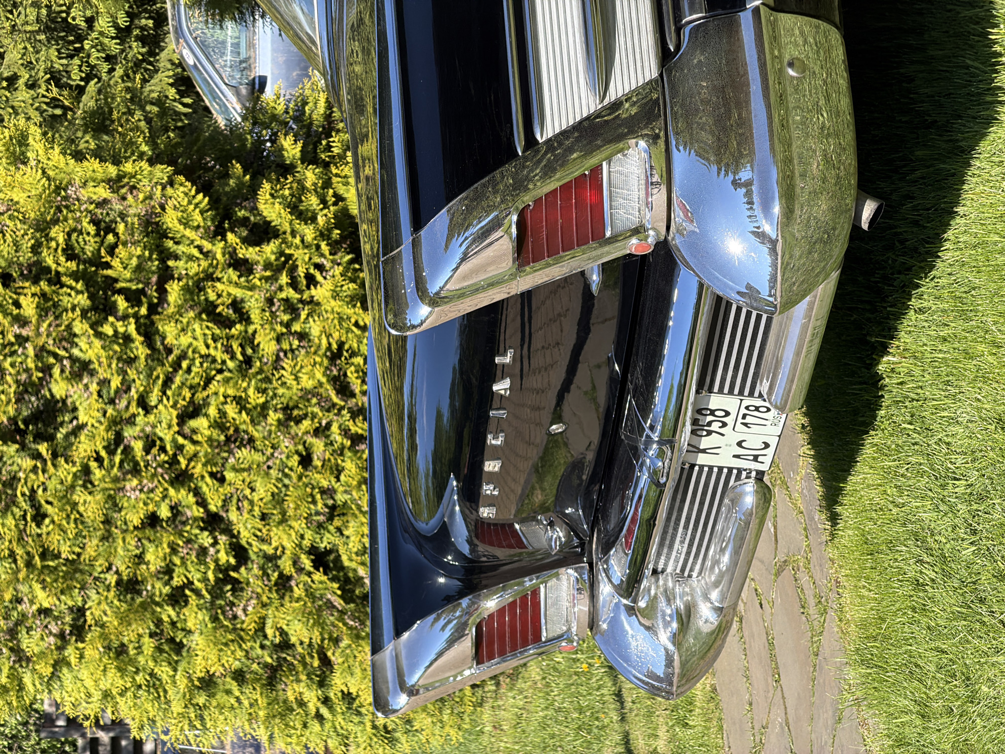 A close-up, rotated image shows the rear of a black vintage car, potentially a classic American model from the late 1950s or early 1960s. The car features prominent chrome accents, including tail fins, taillight housings, and a rear bumper with vertical bars. The car's license plate reads "K 958 AC 178 RUS". The background includes greenery, suggesting the car is parked outdoors. The location is Repino, Russia.