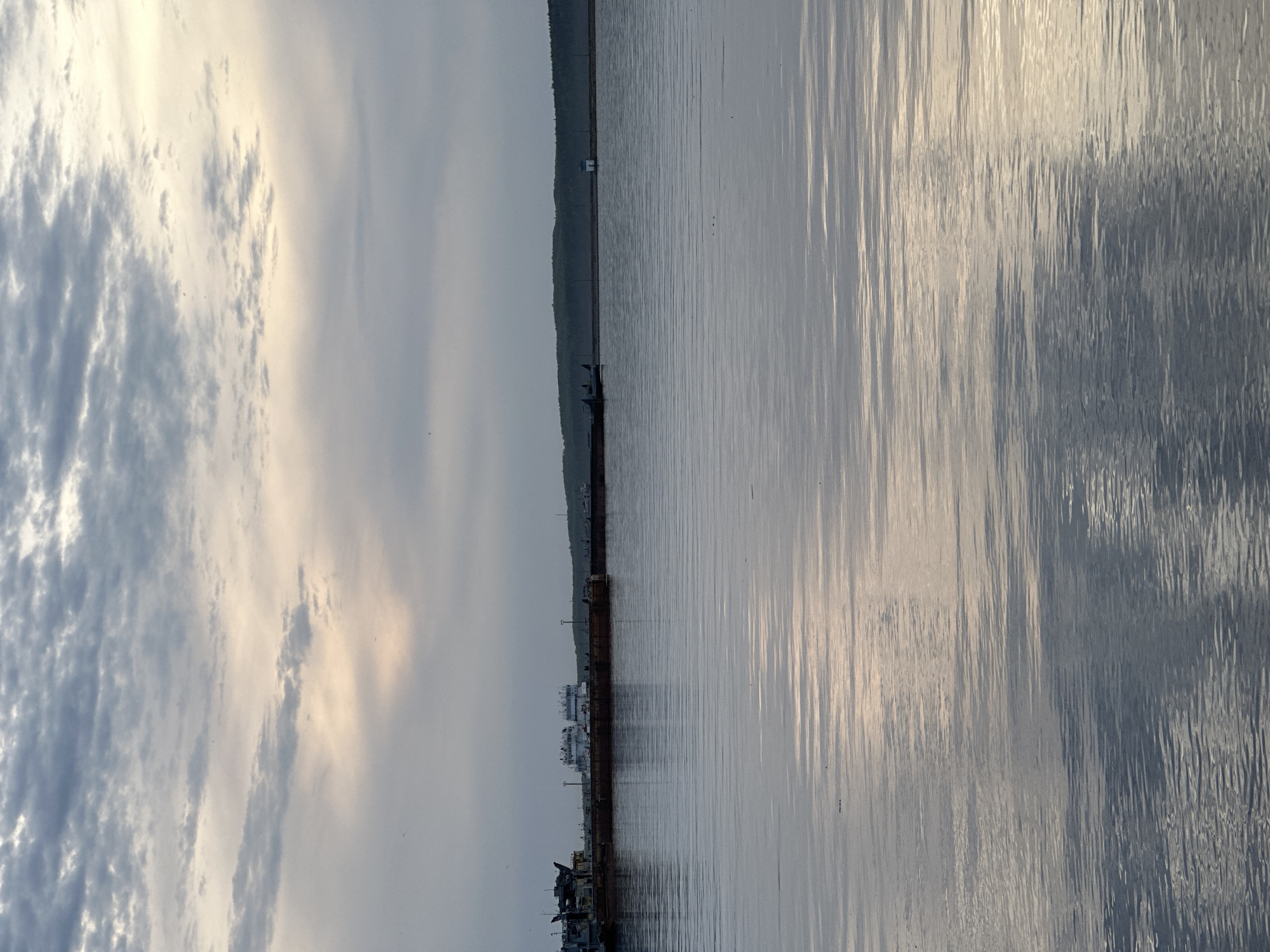 The image depicts a body of water, likely a river or a lake, with an industrial area or dock visible on the opposite shore. The sky is filled with clouds, and the light is subdued, suggesting either early morning or late evening. The water's surface is rippled, reflecting the sky and shoreline. There are boats and buildings visible along the shoreline. The location is inferred to be Chaikovsky, Russia, based on the context.