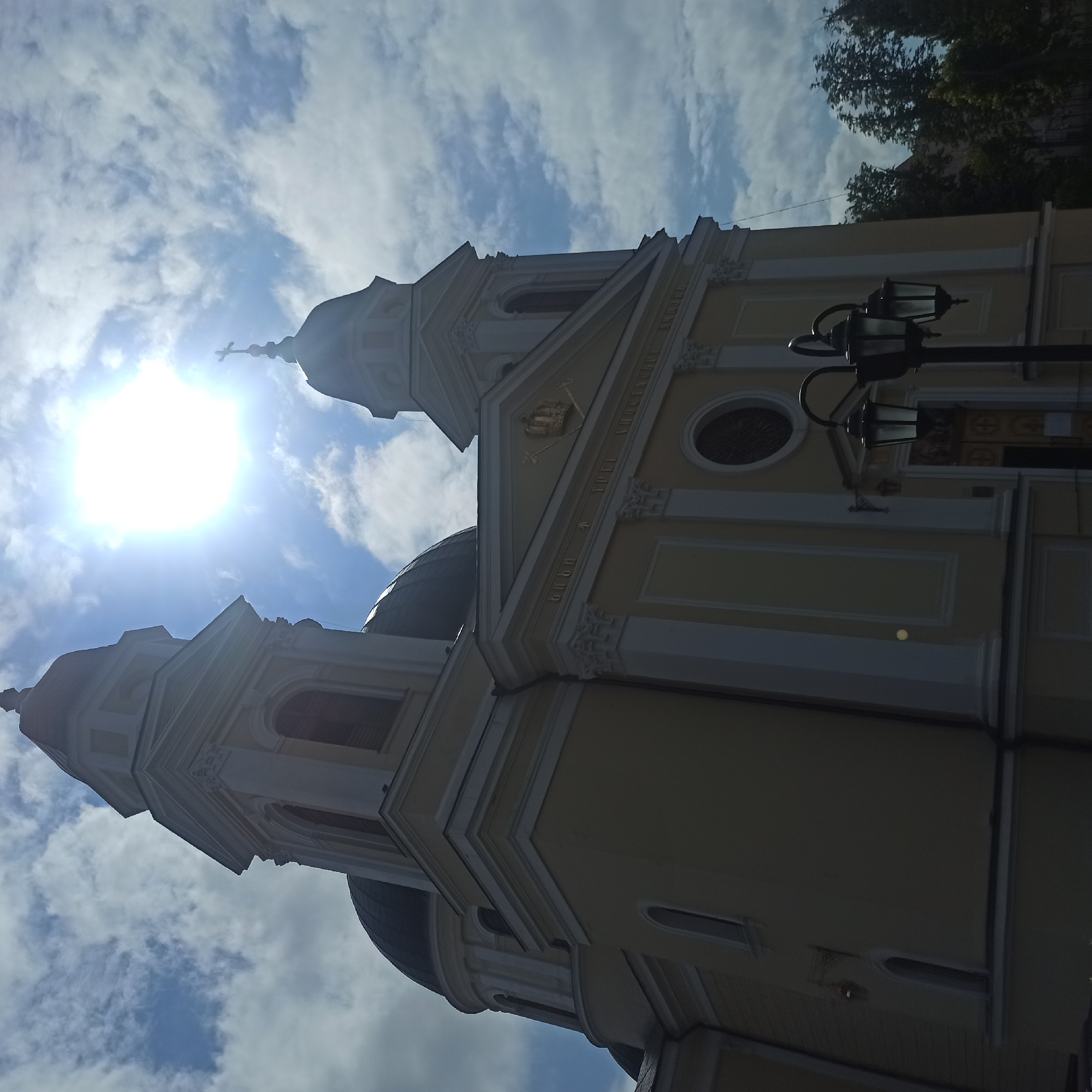 The image shows the Ascension Cathedral in Chernivtsi, Ukraine, viewed from a low angle looking up at the bright sky. The sun is prominently visible, creating a lens flare effect. The cathedral is a light yellow building with dark domes and two bell towers. Architectural details like columns and arched windows are visible. In the foreground, there is a black lamppost with three lights. Trees are visible to the right of the cathedral. The sky is partly cloudy with patches of blue.