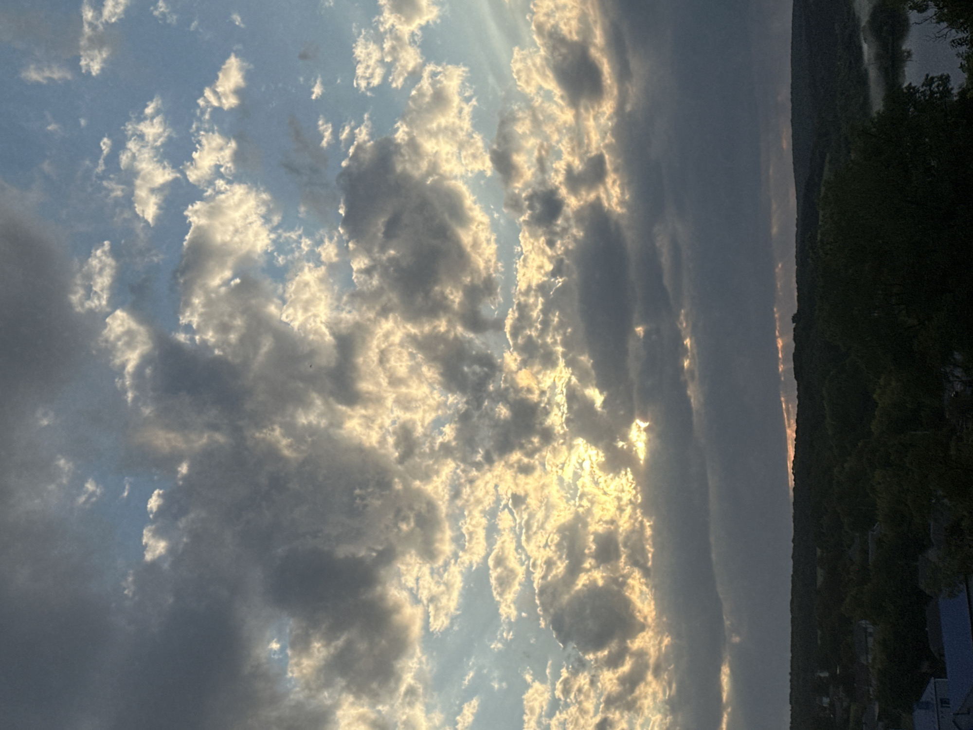 This image depicts a cloudy sky with the sun partially visible through gaps in the clouds, casting a golden glow. Below the sky is a dark, tree-covered landscape with some houses visible. The scene is likely a rural or semi-rural area. The sky takes up the majority of the image, with the landscape occupying the bottom third. The clouds are varied in texture and shade, ranging from dark grey to bright white where illuminated by the sun. There is no discernible action or interaction occurring in the scene. The lighting suggests it is either sunrise or sunset. The location is identified as Makiv, Ukraine, implying this is a view from that area.