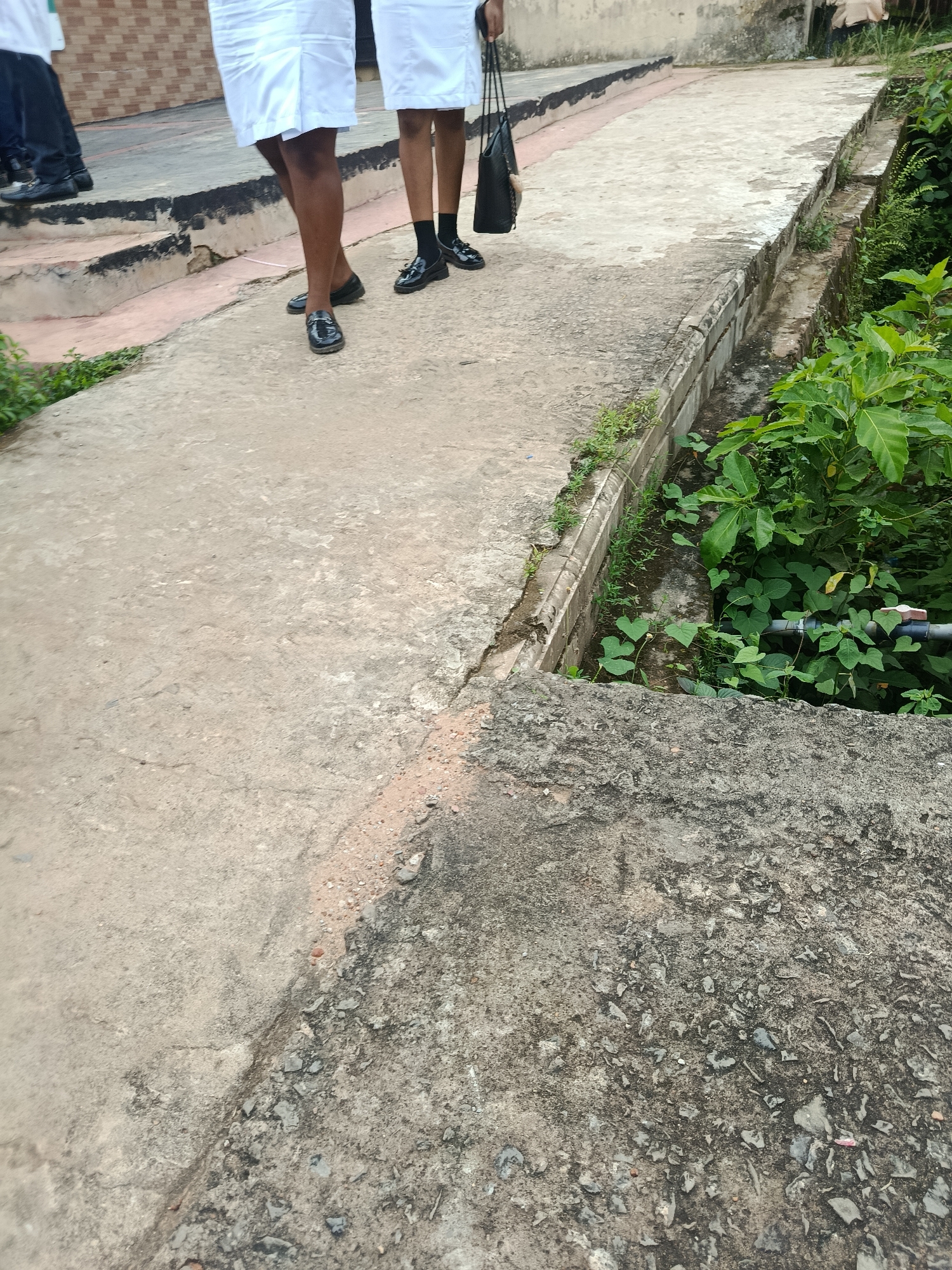 The image shows a partial view of two individuals walking on a concrete pathway in Ukpor, Nigeria. Both individuals are wearing white skirts and black shoes. One person is carrying a black bag. The pathway is bordered by a drainage ditch filled with greenery and a pipe. The ground is rough and uneven, with patches of dirt and stones visible. The background shows a brick building. The scene appears to be outdoors during daylight.