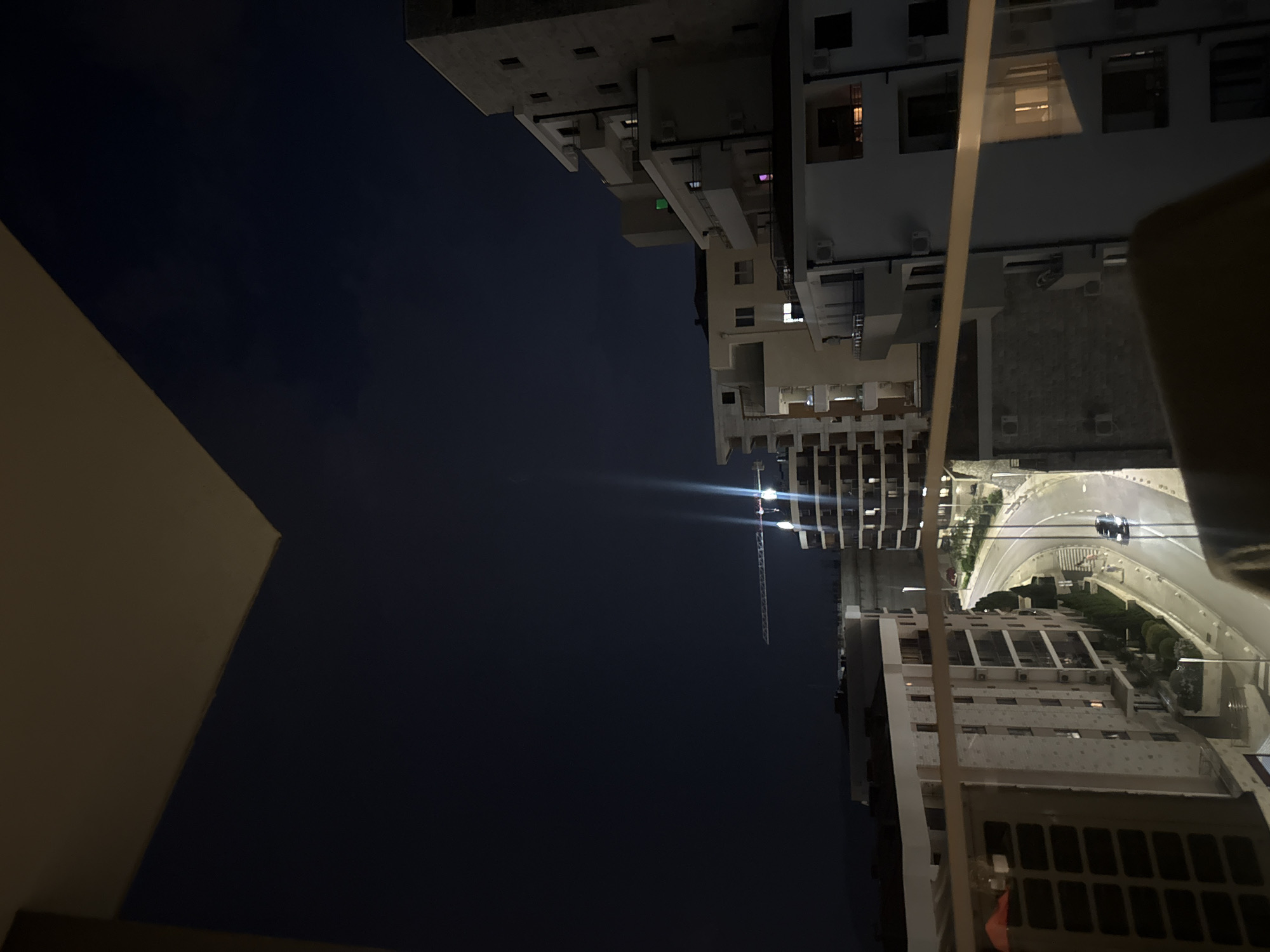 The image shows a nighttime view of a city with several buildings and a road. The perspective is from a high vantage point, looking down and across. The buildings appear to be apartment complexes or office buildings, with many windows visible, some of which are illuminated. One building has a distinct architectural style with many projecting balconies or modular units.
A road curves through the scene, with a single car visible driving on it, its headlights on. Further in the distance, a construction crane is visible, with bright spotlights illuminating its structure. This suggests ongoing construction in the area. The sky is dark, indicating it is nighttime, and there are no visible weather cues like rain or fog.
The scene is urban, with a sense of a developed city. There are no people clearly visible in the image, and the focus is on the architecture and infrastructure. The lighting from the building windows and the streetlights creates a dynamic interplay of light and shadow. The overall mood is one of quiet urban activity at night, with a hint of ongoing development due to the crane. The location cues of Boreti, Montenegro suggest a coastal or Mediterranean setting, though this is not explicitly visible in the image beyond the general impression of a developed urban area. No visible text is present in the image.