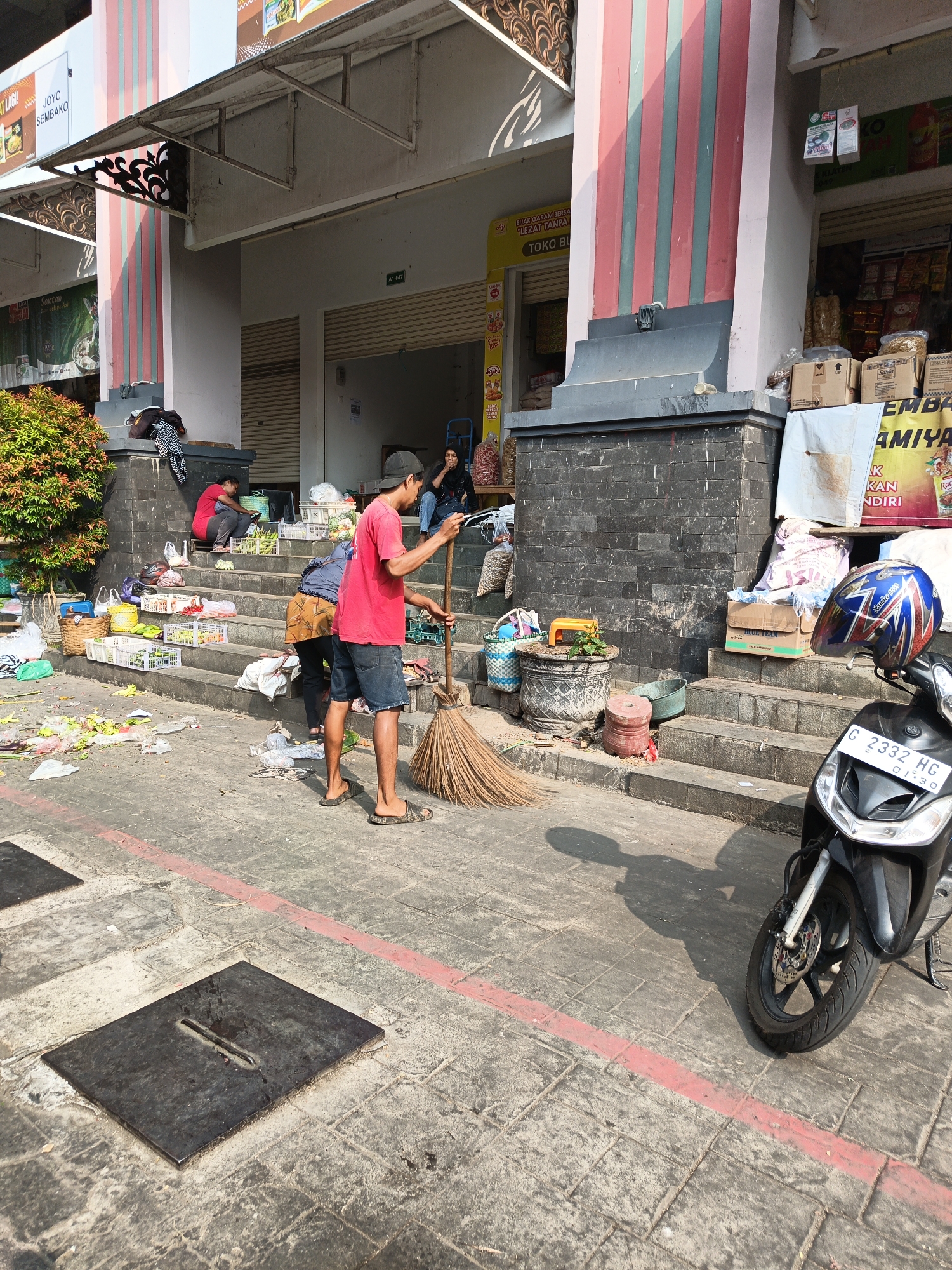 The image depicts an outdoor market scene in Klaten, Indonesia. A young man in a red t-shirt and dark shorts is sweeping the ground with a broom made of dried grass. He is standing on a paved walkway in front of a building with steps leading up to shops. The steps are laden with various goods, including baskets of produce, plastic crates, and bags of what appear to be snacks or groceries. Debris, including discarded food items and plastic bags, is scattered on the ground and steps, suggesting a recent market day or a general lack of tidiness. Several people are visible in the background, some seated on the steps and others standing near the shops. A scooter is parked on the right side of the image, with a helmet resting on its handlebars. The building's facade features red and green striped columns and a dark stone base. The overall impression is a bustling, yet somewhat unkempt, marketplace environment.