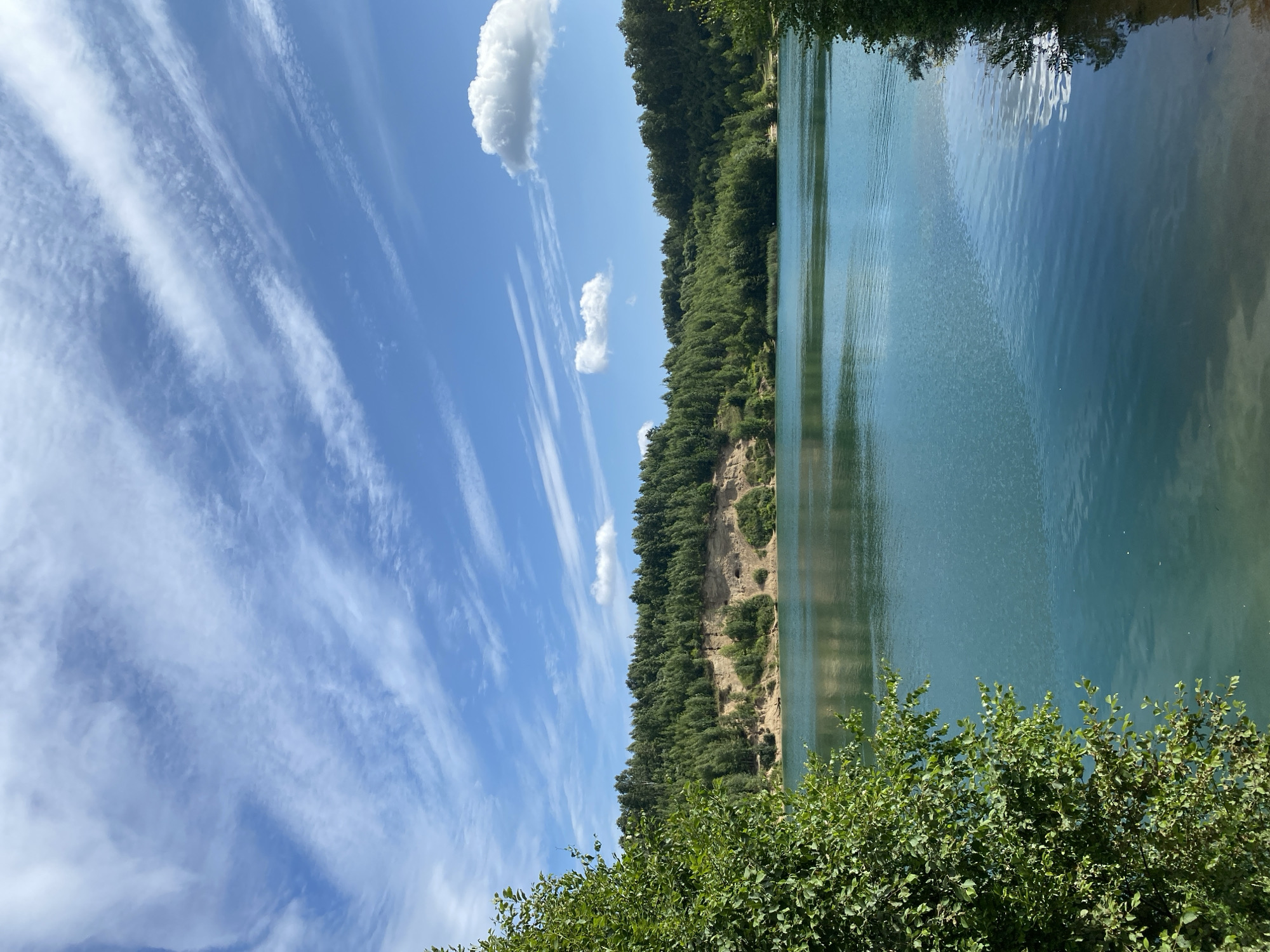 A vertical, eye-level, medium shot captures a serene natural landscape under a bright, partly cloudy sky. The foreground features lush green foliage framing the left and right edges of the scene, with the left side showing more abundant growth. The mid-ground is dominated by a tranquil, turquoise lake, its surface gently rippling. The lake is bordered by a sandy cliff face, dotted with patches of greenery and small caves, which gives way to a dense forest of green trees stretching across the horizon. The sky above is a vibrant blue, adorned with wispy white clouds that streak across the expanse, with a few larger, puffy clouds also present. The overall impression is one of a picturesque, natural retreat, likely a quarry or lake formed in a former mining area.