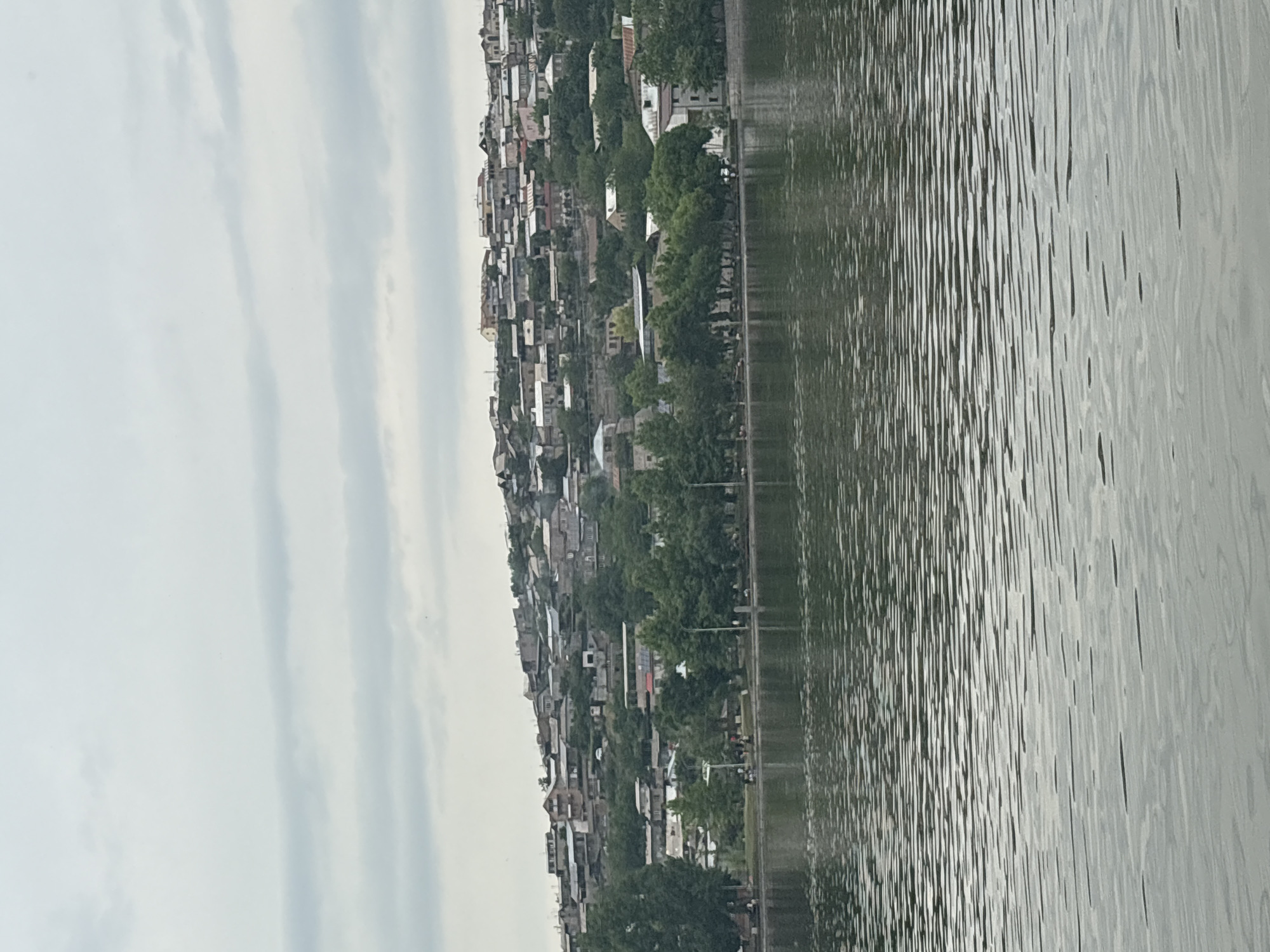 The image depicts the periphery of Yerevan Lake in Armenia, viewed from the water's edge. On the opposite shore, a dense collection of residential buildings climbs the hillside, interspersed with green trees. A line of trees follows the shoreline. The lake itself occupies the foreground, its surface rippled with wave patterns. The sky above is overcast, with pale grey clouds stretching across the horizon. There is a waterside promenade.