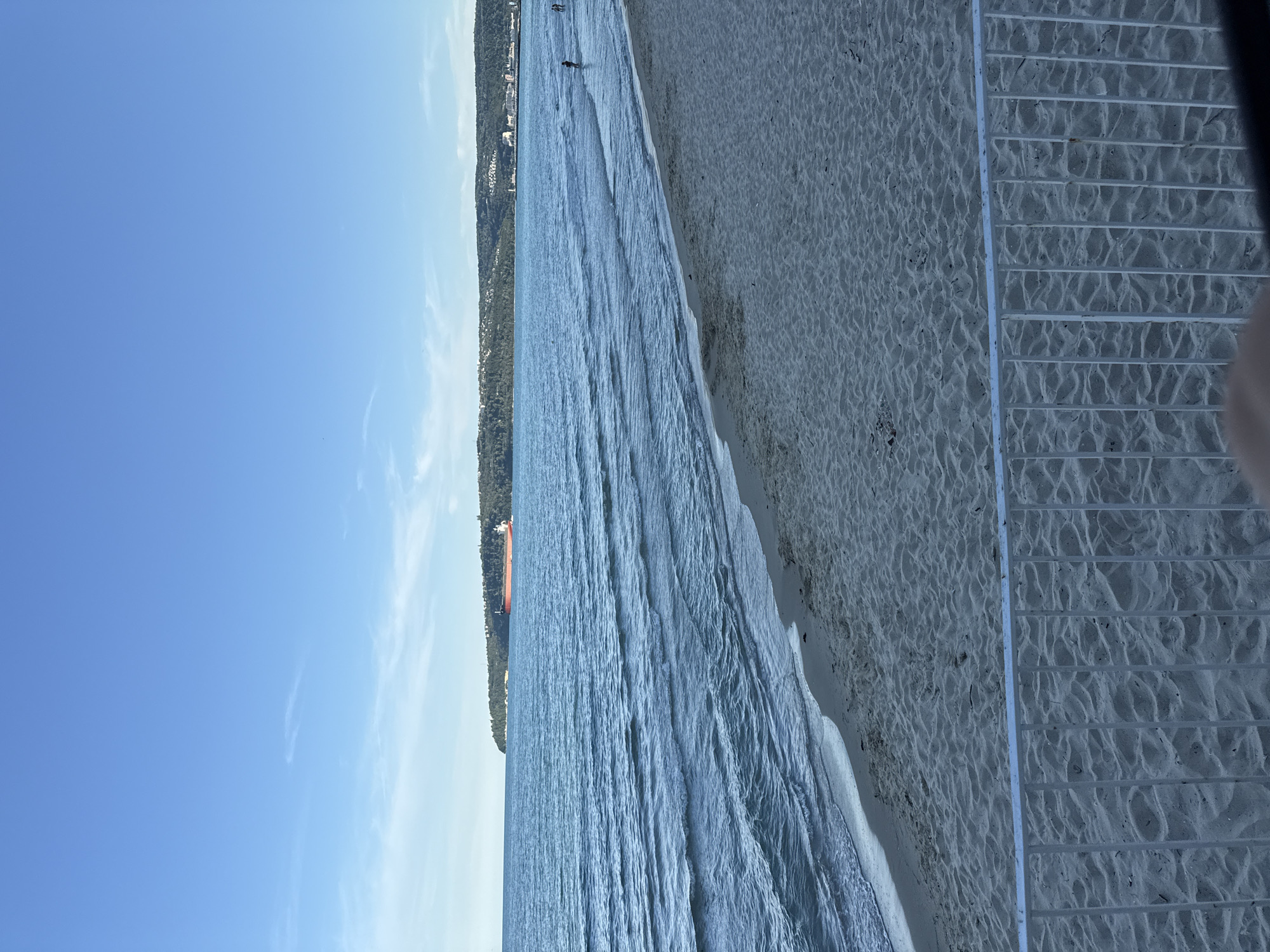 The image depicts a wide view of a sandy beach with gentle waves lapping at the shore under a clear blue sky. In the distance, a large, red cargo ship is visible on the horizon, alongside a tree-covered coastline with scattered buildings. A few people can be seen wading in the shallow water near the shore. A white metal railing runs along the bottom of the frame, partially obscuring the sand. The overall scene suggests a peaceful day at the beach.