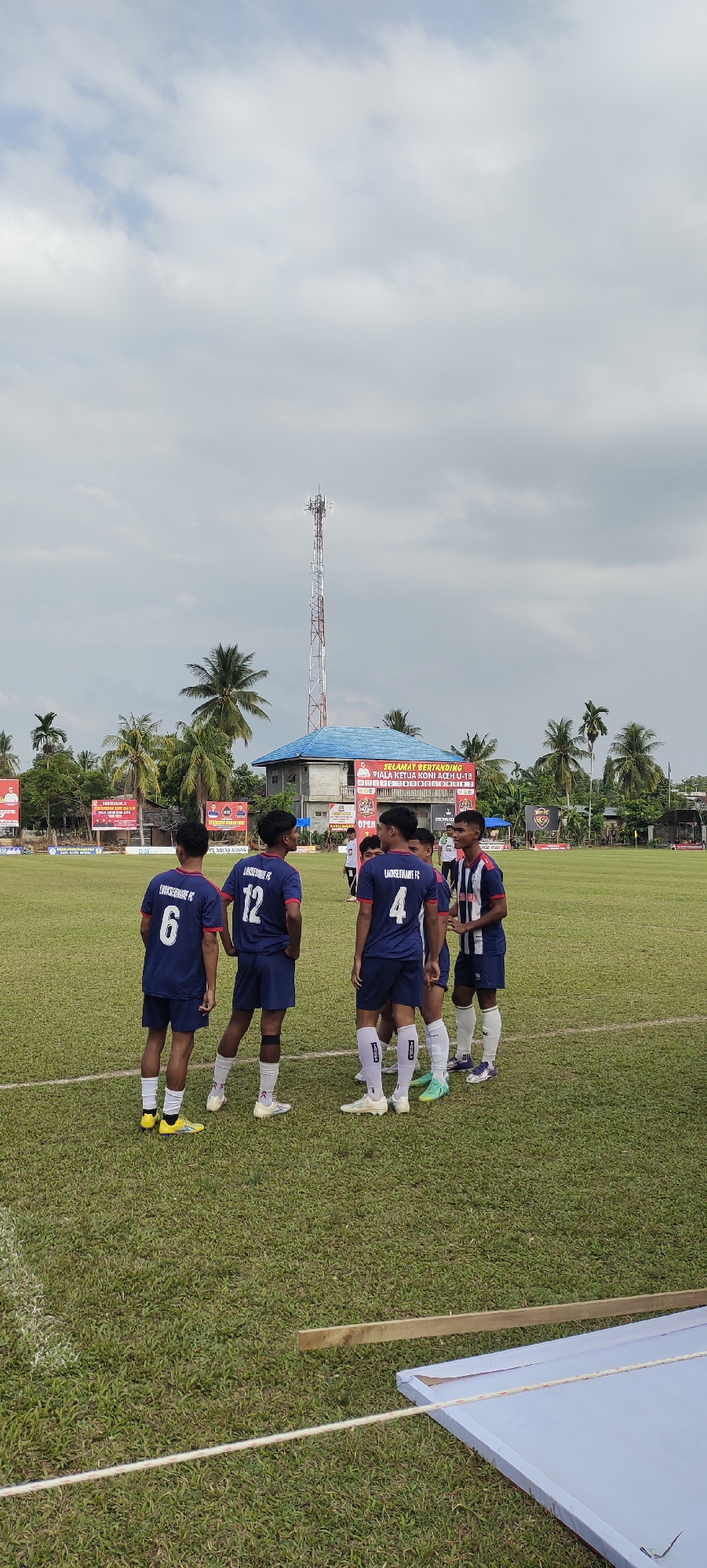 This is a vertical, full-shot, eye-level photograph of a group of young male soccer players in navy blue uniforms with white numbers on the field in Kilometer Delapan, Indonesia. The players are gathered in small groups on a bright green grass field, with white lines marking boundaries. The sky is cloudy and overcast, with a tall cellular tower visible in the background behind a low-rise building with a blue roof. There are palm trees and other vegetation surrounding the field, and several banners are displayed in the background.
In the foreground, four players are standing facing towards the right. The player on the far left wears jersey number 6, the next player wears number 12, and the player to his right wears number 4. They are all wearing navy blue jerseys and shorts with white socks. The player on the far right is wearing a different navy blue and white striped jersey.
A bit further back, to the right of the main group, are three more players in navy blue jerseys. One of them appears to be interacting with a player from the other team.
The environment suggests a soccer match or practice is taking place. The overall lighting is diffused, consistent with an overcast sky. The scene is outdoors, on a sports field.