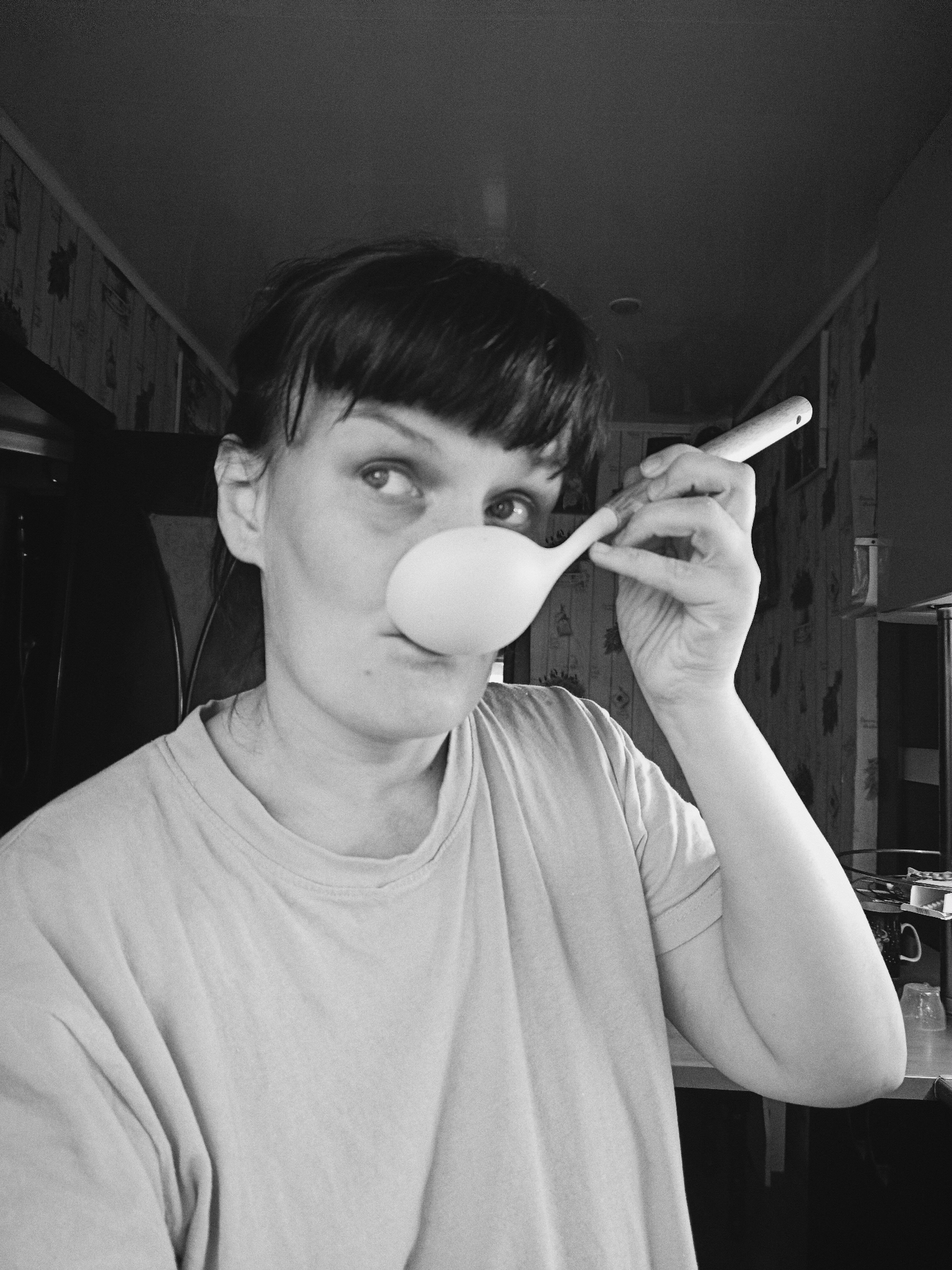 This black and white, grainy photograph features a woman playfully balancing a large white spoon or ladle on her nose. She is looking upwards and to the right with a concentrated yet whimsical expression, her mouth slightly pursed. Her dark, short hair has bangs across her forehead. She is wearing a light-colored, round-necked t-shirt. Her right hand gently supports the handle of the spoon.
The background of the image reveals an indoor setting, likely a home. The walls are covered with patterned wallpaper featuring a repeating floral or leaf design. A ceiling is visible in the upper part of the frame, and to the right, a countertop or table holds a clear glass and other indistinct objects, suggesting a kitchen or a casual domestic space. The overall mood of the image is candid and lighthearted. No text is visible in the photograph. The location is Фоки, Russia.