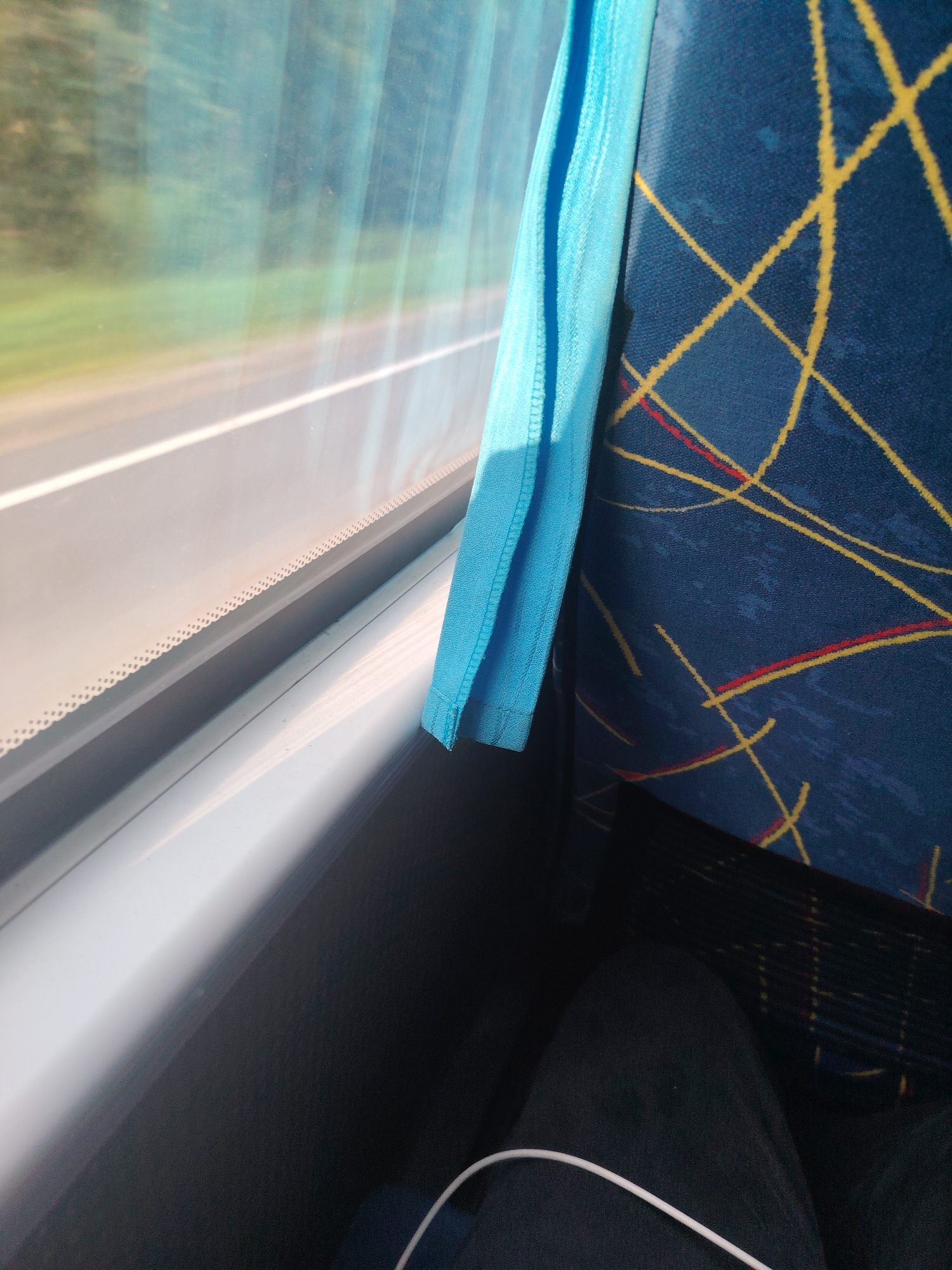The image depicts the interior of a vehicle, likely a bus, with a view through the window. A bright blue fabric hangs down on the right side of the window. The window shows a blurred landscape of greenery and a road with a white line, indicating movement. The interior features a seat upholstered in dark blue fabric with a geometric pattern of yellow and red lines. A portion of a person's dark clothing and a white cable are visible in the lower right corner. The lighting suggests it is daytime. The provided location of Shetakovo, Russia, is noted but not visually verifiable from the image itself.