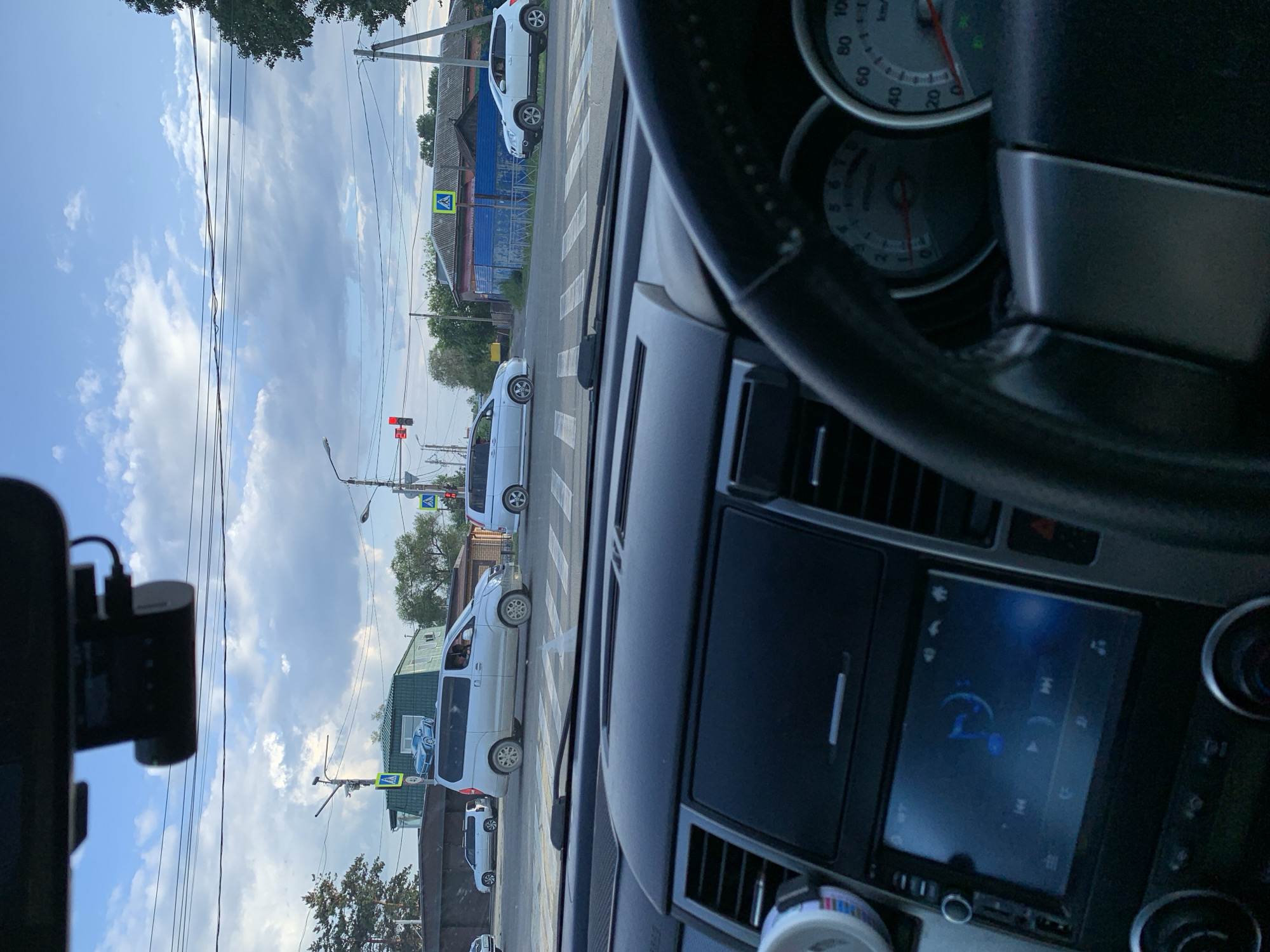 The image is taken from inside a vehicle, looking forward through the windshield. A dashboard, steering wheel, and instrument cluster are visible in the foreground. Outside the vehicle, a street intersection with traffic lights and crosswalks is depicted. Several vehicles are present, including a silver minivan, a white crossover SUV, and another silver minivan. Pedestrian crossing signs are visible on poles. Buildings and trees line the street, and the sky is cloudy. A dashcam is mounted on the windshield. The scene is consistent with a traffic situation in a city, possibly Belogorsk, Russia, given the information.