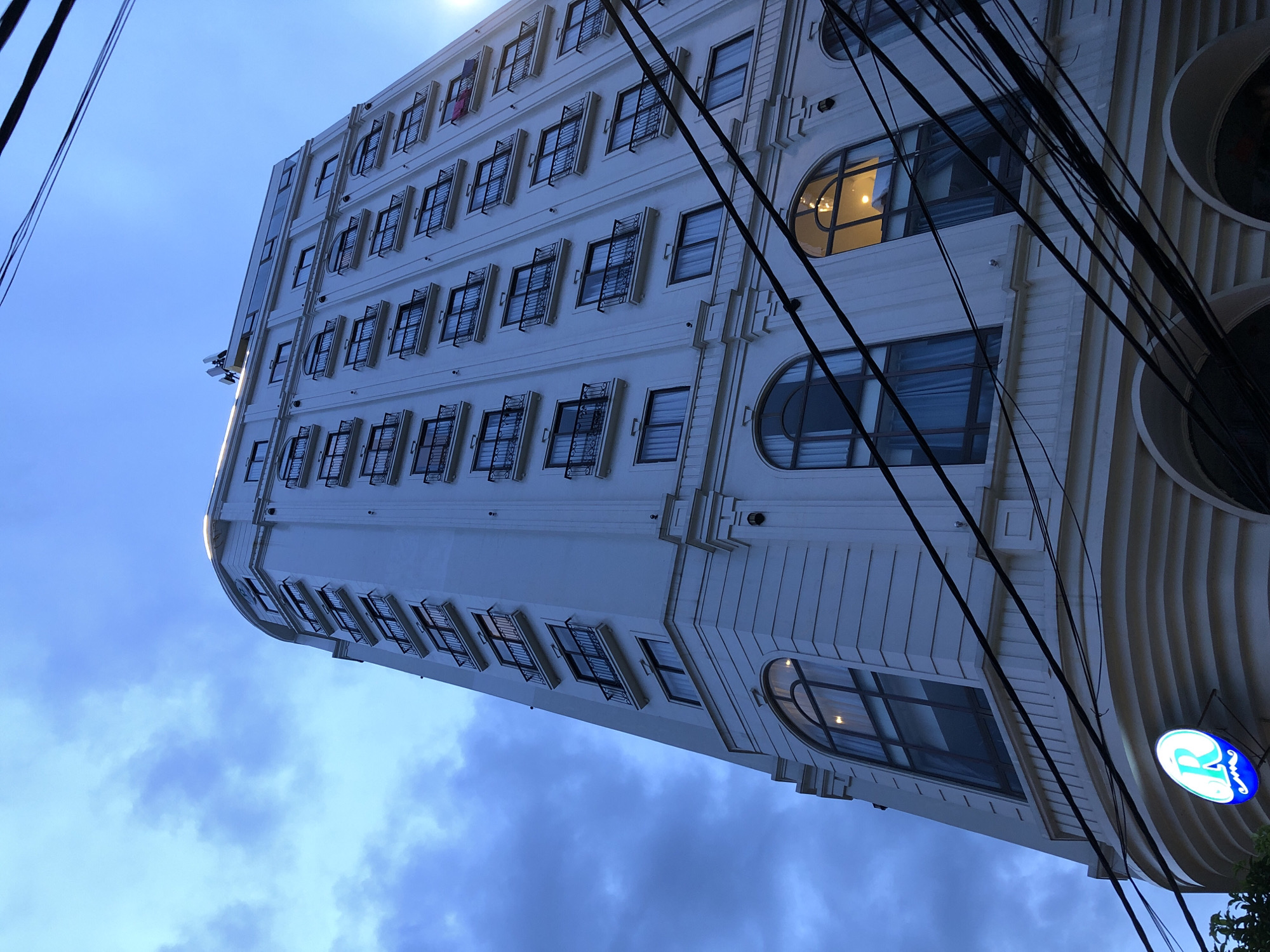 This is a photograph taken from a low angle, looking up at the exterior of a modern, white, multi-story building in Đà Nẵng, Vietnam. The building has a curved facade and is adorned with numerous windows, many of which are arched on the lower levels. Each window on the upper floors appears to have a small balcony with black railings. The sky above is a cloudy, pale blue, indicative of either early morning or late afternoon. A prominent feature of the building's base is a circular blue sign with a white outline and the letter 'R' inside, accompanied by a decorative swoosh and what appears to be a mustache-like element. A dense network of dark electrical wires crisscrosses the foreground, obscuring parts of the building. The overall impression is urban and contemporary, with the building being the primary subject against a backdrop of the sky and overhead utilities.