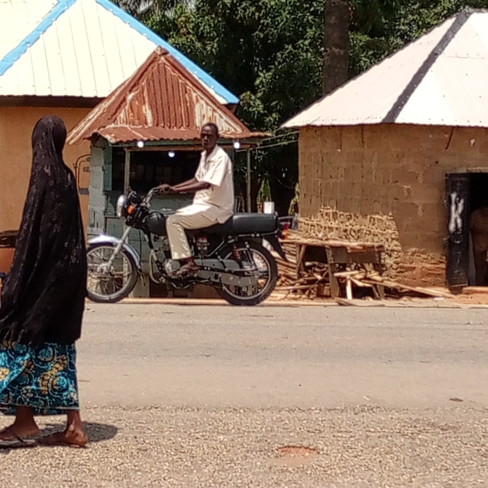 A daytime, eye-level shot depicts a street scene in Mararaba, Nigeria. A woman in a dark head covering and patterned blue and gold skirt walks left to right on the left side of the frame. To the right of the woman is a man wearing a light-colored shirt and pants, sitting on a black motorcycle that faces left. He appears to be looking at something off-camera. Behind the man and motorcycle is a small structure with a rusty corrugated metal roof and a blue-painted facade. To the right of the motorcycle and further back is a building constructed of mud or similar material with a white "K" painted on the doorframe. Wood is stacked or scattered near the building. The road runs horizontally across the midground. The foreground is gravel or a similar material. Trees are visible in the background.