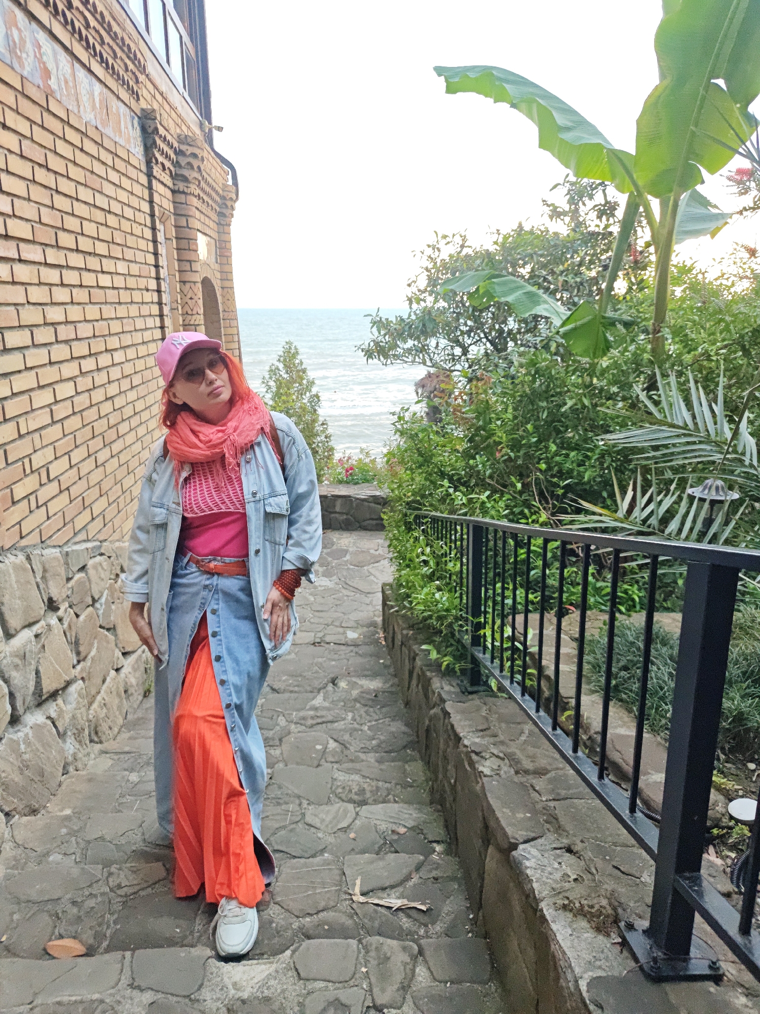 A woman stands on stone steps next to a building constructed with a mix of brick and stone. She is wearing a pink baseball cap, orange sunglasses, a peach-colored scarf, and a denim jacket over a pink top. Her skirt is a combination of denim and an orange pleated fabric. White sneakers adorn her feet.
To her right is a metal railing with green foliage behind it, including a banana tree. In the background, the ocean can be seen under an overcast sky.
The location is in Sochi, Russia.