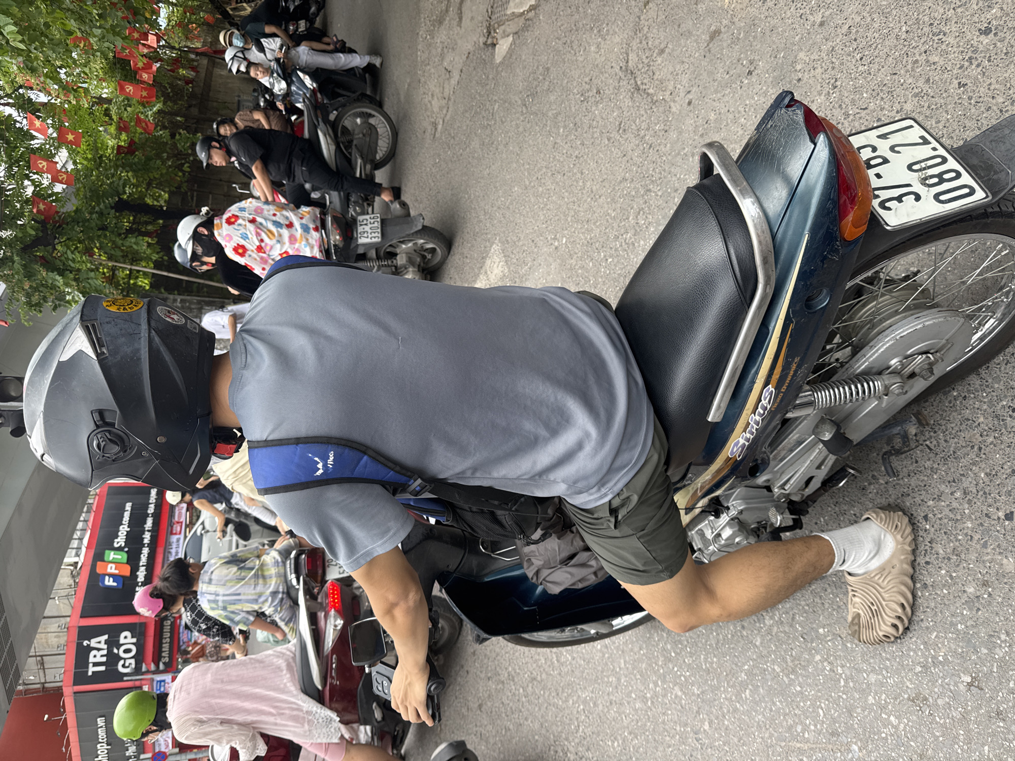 A man wearing a gray t-shirt, olive green shorts, white socks, and beige sandals rides a dark teal motorcycle in a street scene in Hanoi, Vietnam. He is wearing a black helmet. The motorcycle's license plate reads "37-B5 080.21". In the background, other motorcycles and people are visible, as well as a red building with signage that appears to be an FPT Shop. Red flags with yellow stars hang from trees overhead.