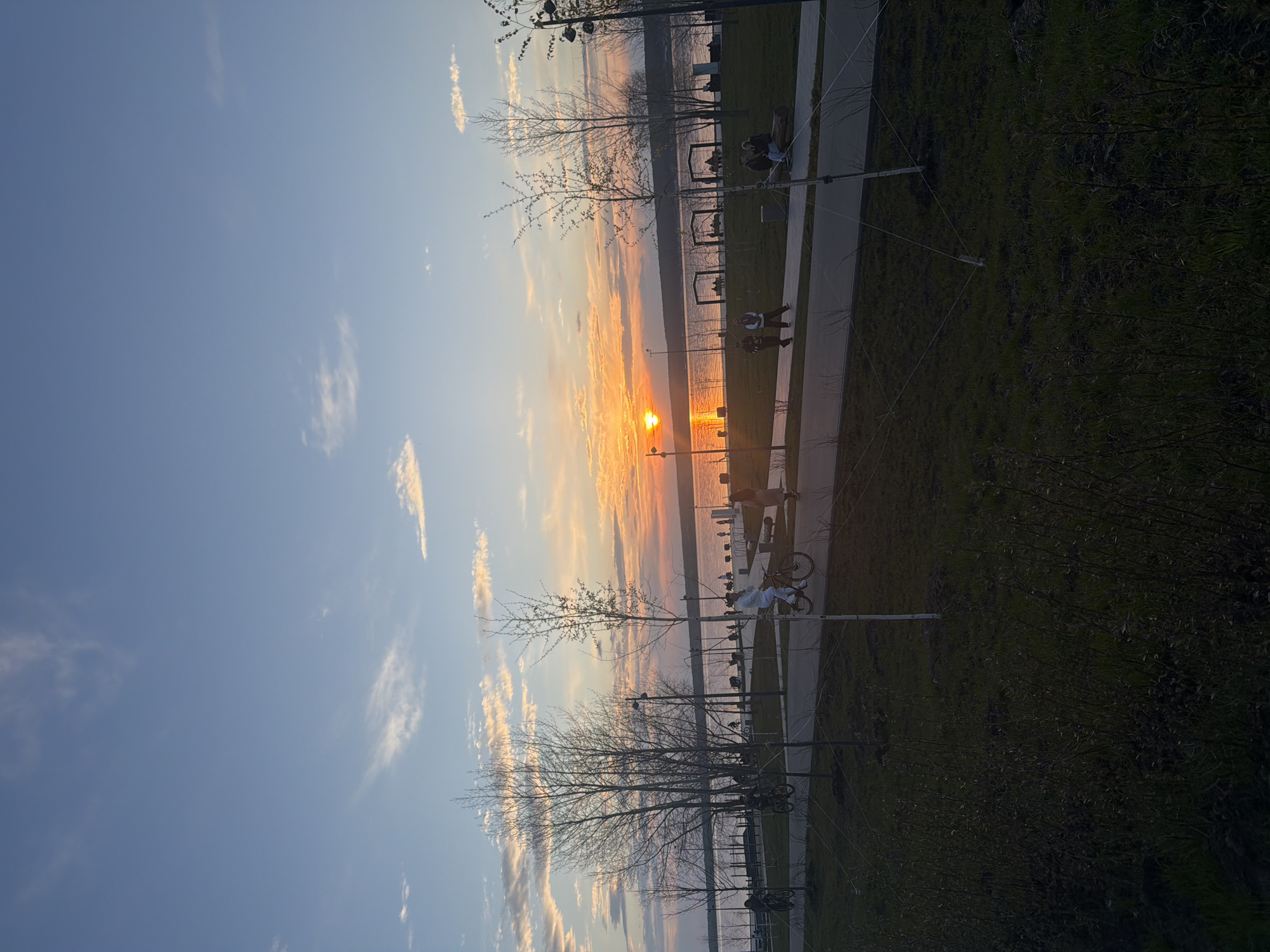 Captured in Naberezhnye Chelny, Russia, the image portrays a sunset over a body of water. The sun's position suggests it is near the horizon, with its light reflecting off the water surface. The foreground features trees with bare branches, indicating a seasonal time frame like late fall or early spring. A paved path runs alongside the water's edge, with people walking and sitting on benches. Bicycles are also visible along the path. The sky is a mix of blue and orange hues, indicative of the sunset. Vegetation is seen on the right side of the path.