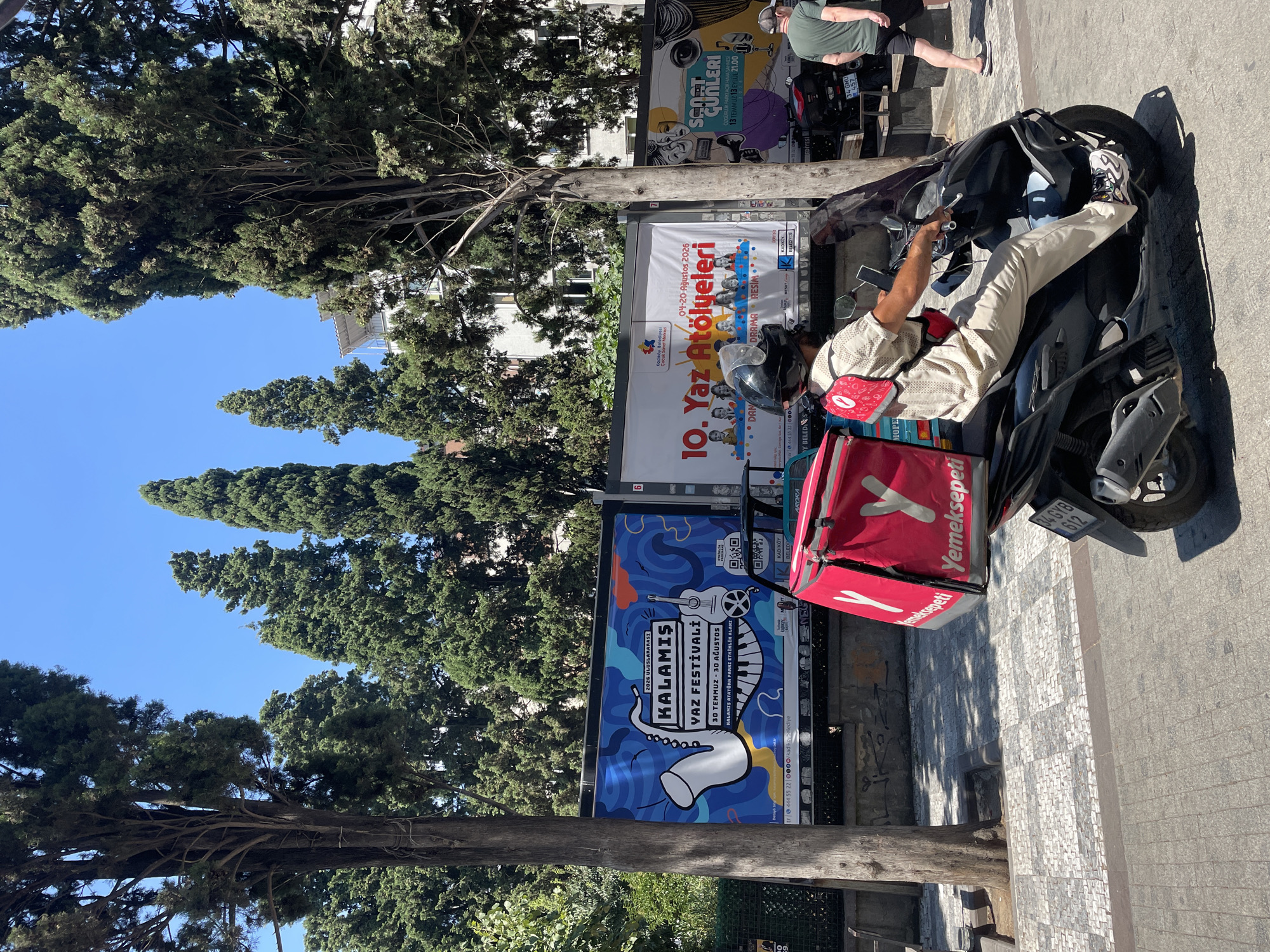 A delivery person on a scooter, wearing a light-colored uniform and a helmet, transports a large, red insulated bag bearing the "Yemeksepeti" logo. The scooter has a license plate reading "34 GHY 612". The delivery person is riding on a street in Kadıköy, Turkey, with a sidewalk paved with a checkered pattern of light and dark tiles. Behind the scooter, large billboards display event advertisements, including one for "KALAMIŞ YAZ FESTİVALİ" (Kalamış Summer Festival) and another for "10. YAZ ATÖLYELERİ" (10th Summer Workshops). Tall cypress trees line the street, and a white building is visible in the background. A second scooter and another person, wearing a green t-shirt and dark shorts, are also present on the right side of the image. The sky is clear and blue.