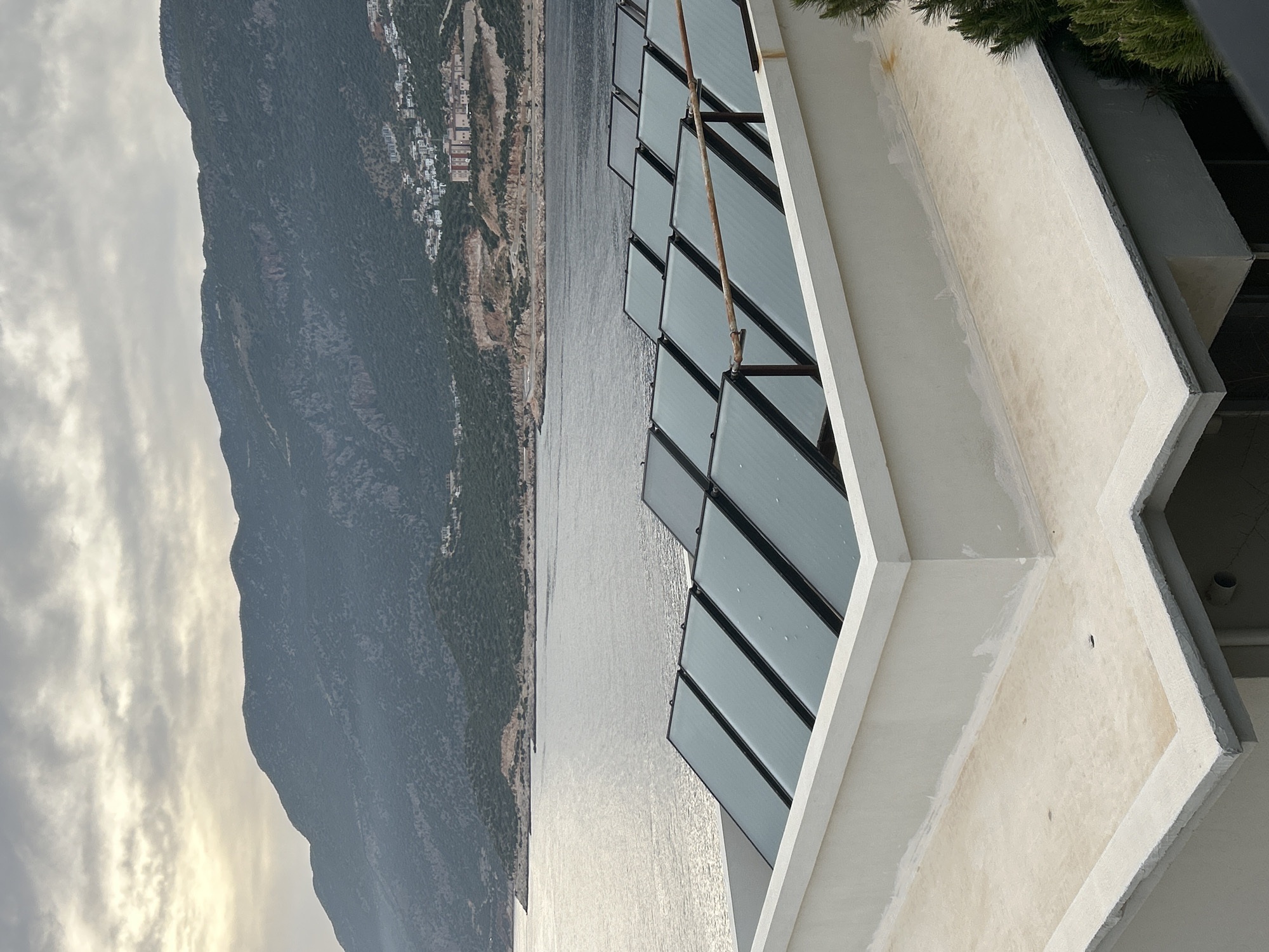 The image captures a coastal scene in Kaş, Turkey. A mountain with a city is seen in the background across a body of water. A building with a series of windows is on the right side of the photo. The sky is cloudy.