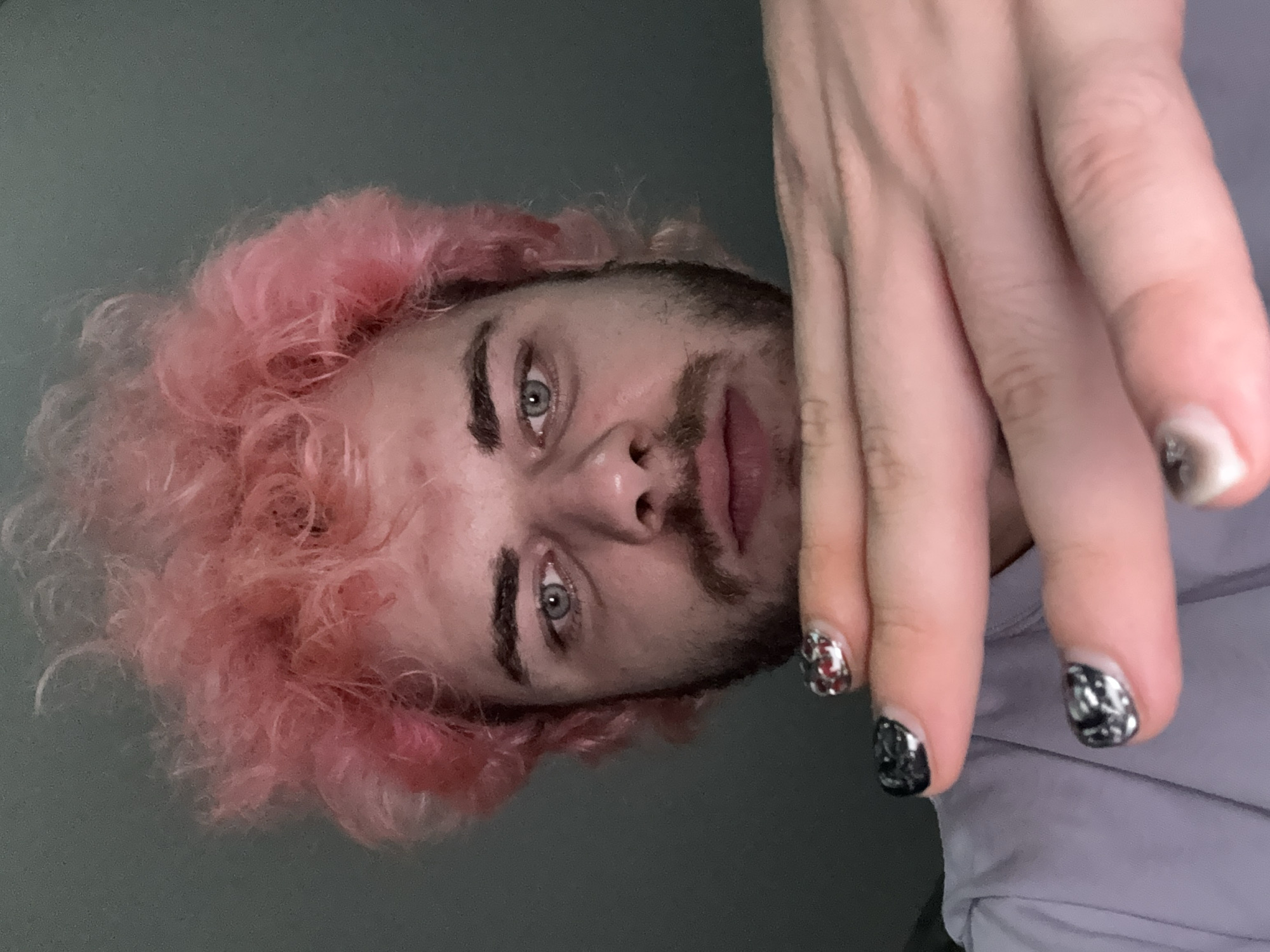 The image shows a person with pink, curly hair and a short beard. They have light skin and light-colored eyes. Their hand is raised in the foreground, obscuring part of their face. The fingernails on the hand are decorated with silver and red polish. The person is wearing a light purple shirt or jacket. The background is a plain grey. There is no indication of the location being Đà Nẵng, Vietnam.