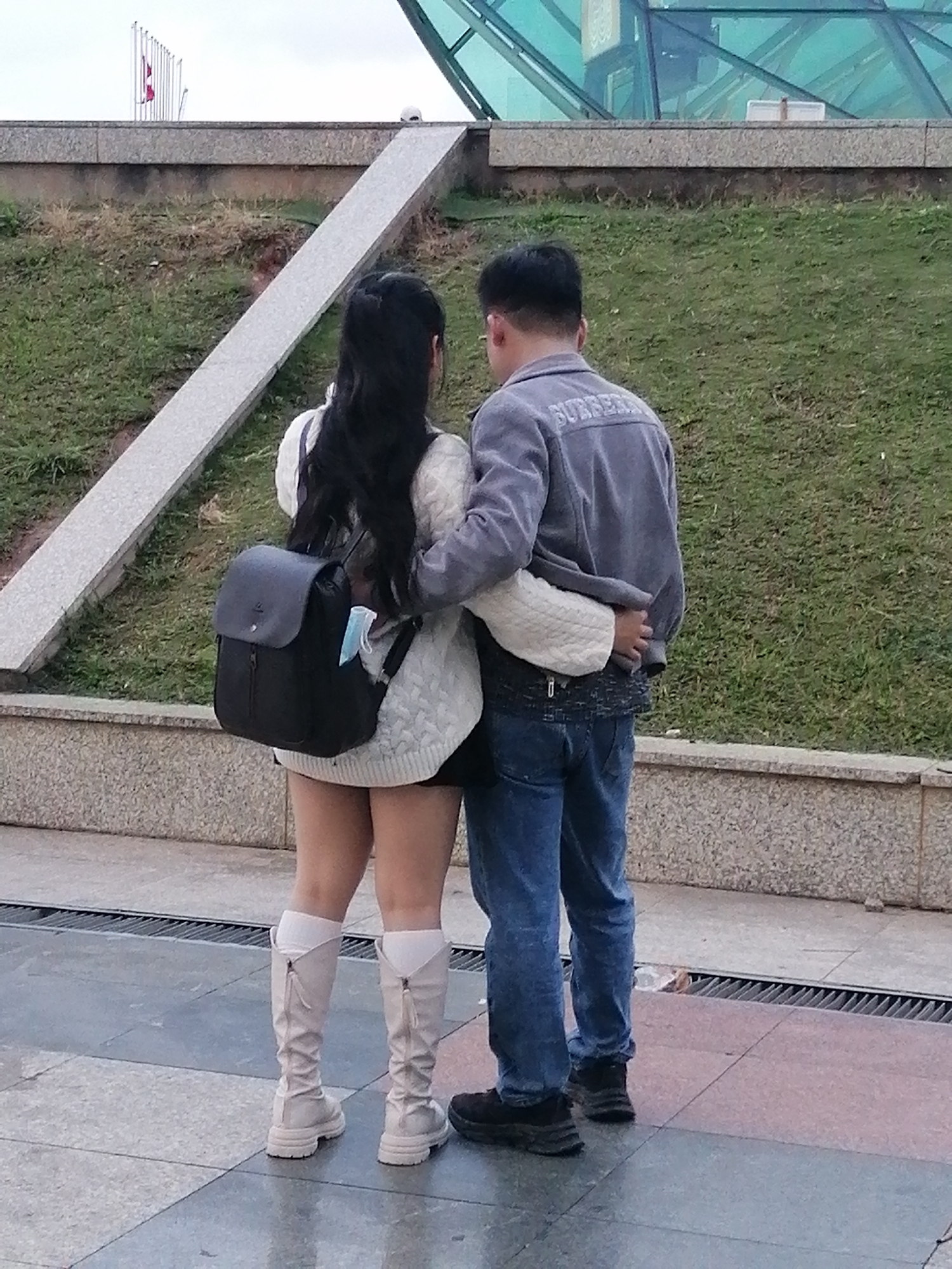 A young couple, a man and a woman, stand facing away from the camera, embracing. The woman has long black hair and wears a white cable-knit sweater and a black skirt. She also has on white, knee-high boots with zippers and what appears to be a black backpack with a mask hanging from it. The man wears a grey jacket with "BURBERRY" printed on the back and blue jeans. They are standing on a paved area with a patterned floor. Behind them is a grassy slope with a grey concrete pathway leading upwards. Further in the background, there is a modern glass structure and a series of flagpoles. The overall environment suggests an outdoor public space in Da Lat, Vietnam, likely a park or urban plaza.