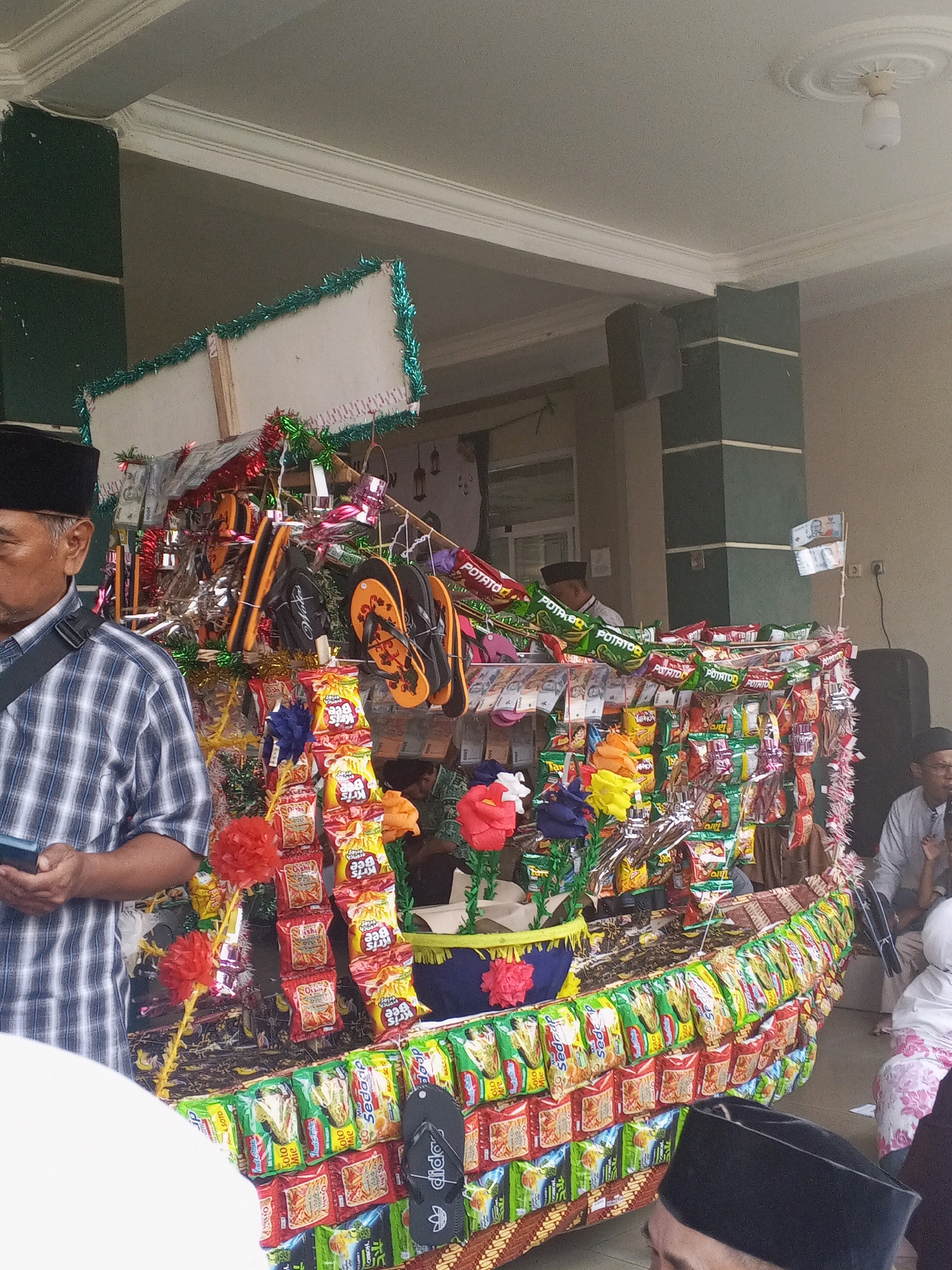 A man wearing a plaid shirt and a black peci hat stands on the left side of the image, holding a phone. In front of him is a decorated float, shaped like a boat, adorned with snacks and other items. The float is covered in colorful snacks, such as potato chips and instant noodles, and also features flip-flops, artificial flowers, and what appear to be paper money. The float is decorated with green and red tinsel. In the background, other people are visible, including a person wearing a white headscarf and a person in a light grey shirt. The scene appears to be outdoors, possibly at a street event or festival. The architecture in the background suggests a building with pillars and a ceiling with decorative molding, consistent with structures found in Ciruas, Indonesia.
