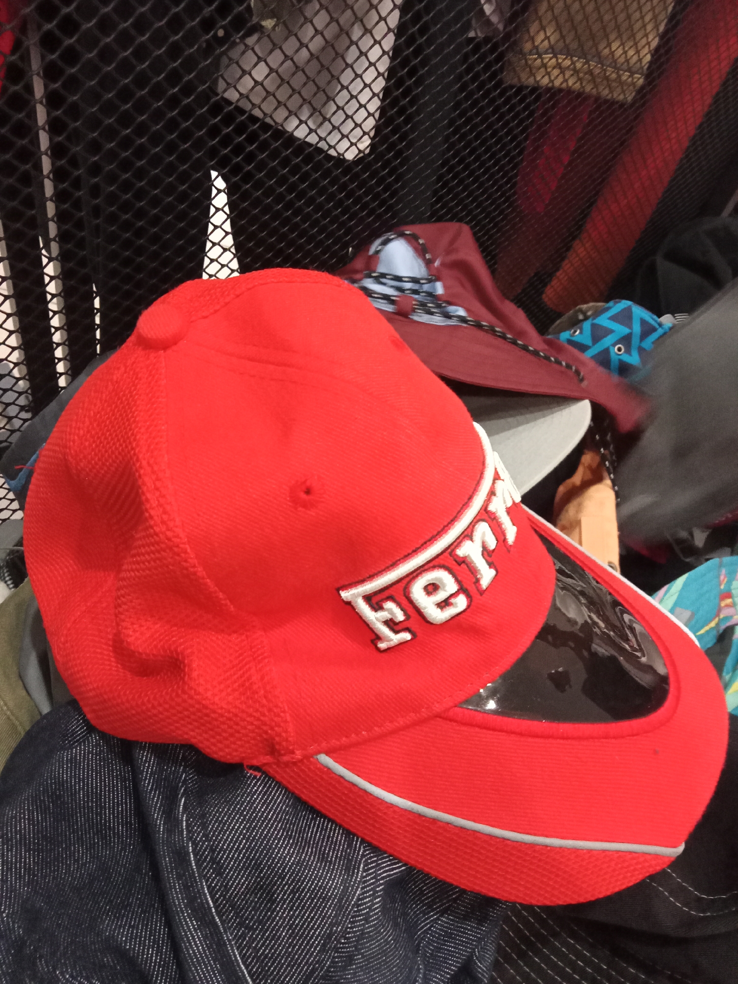 A red baseball cap with "Ferrari" embroidered in white on the front is displayed prominently in the foreground, angled with its brim facing the viewer. The cap appears to be made of a textured fabric, possibly with a mesh-like pattern on the sides. A clear visor is attached to the brim, reflecting the surroundings. The cap is resting on a dark, possibly denim, fabric. Behind the cap, a black mesh grate is visible, suggesting it might be part of a storage unit or a barrier. Various other objects, including what appear to be other hats or folded fabric in shades of red, blue, and gray, are visible behind the main subject. The overall environment seems to be indoors, with a cluttered arrangement of items. The lighting suggests an indoor setting with artificial light. The image does not provide information about the specific city or location.