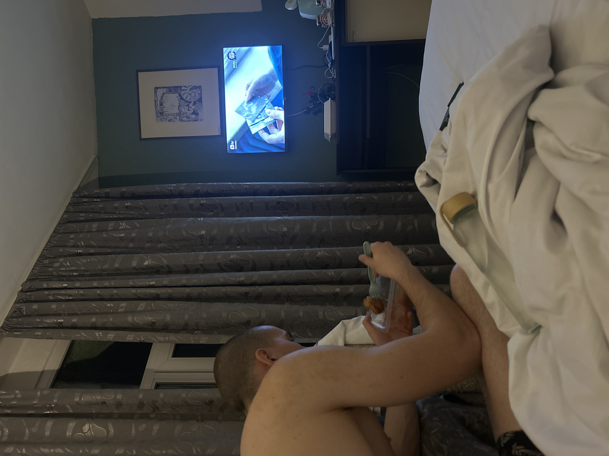 The image depicts an indoor scene, identified as a hotel room in Varna, Bulgaria, due to the "bTV" channel logo and the text "ВАРНА 22:00" visible on a wall-mounted flat-screen television. A male individual, shirtless with short hair, is seated in the mid-ground, observed from the back and right side, actively consuming food from a clear plastic container using a light blue plastic fork. A second individual's upper body, clothed in white, is partially visible beneath the primary subject's right arm. The room features a dark teal accent wall with a framed print depicting a stylized face positioned above the television. Below the television, a dark brown console holds a white takeaway box lid, a small black fan, and various cables. A white mini-fridge is partially visible to the right of the console. Dark grey patterned curtains cover a window on the left side of the room. In the foreground, a bed with white linen is visible, upon which a clear water bottle with a yellow cap and a black rectangular object are resting.