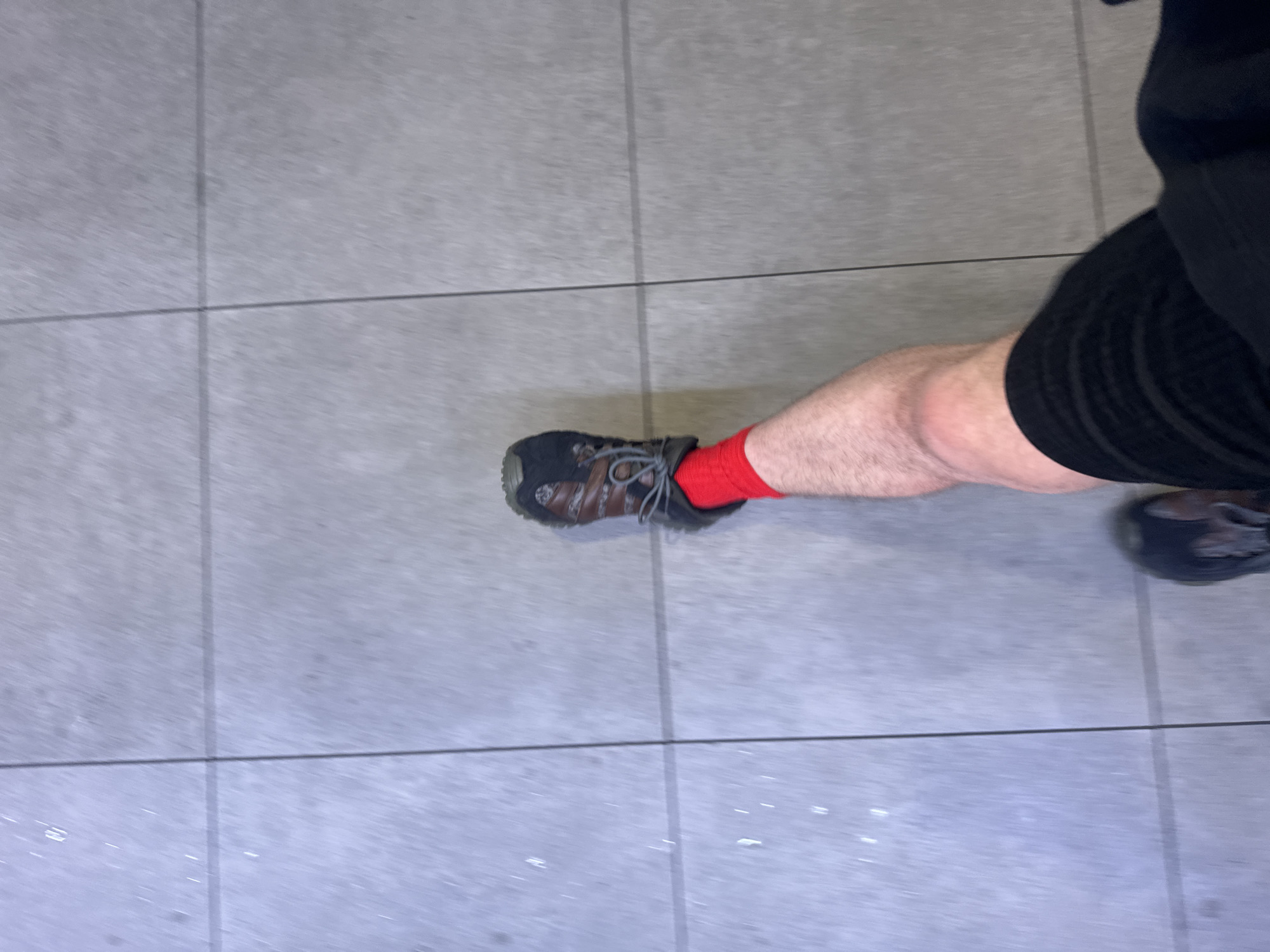 A high-angle, slightly blurry photograph shows a person standing on a tiled surface. The person is wearing black shorts, red socks, and dark hiking shoes. The surface appears to be light grey tiles with dark grout lines. The scene suggests an indoor setting, possibly a public space. There is no information to determine its location in Uttlesford, United Kingdom.