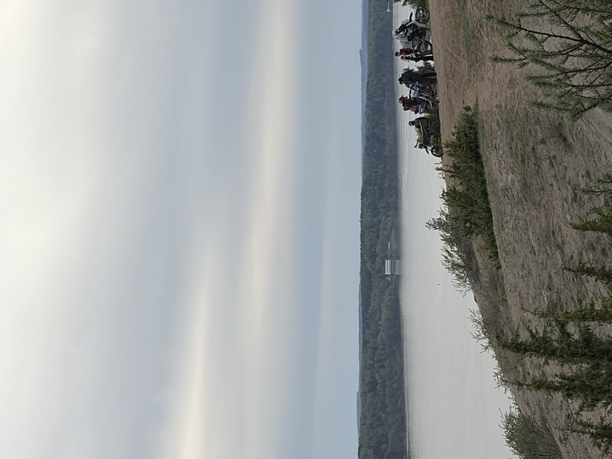 This image shows a body of water in Ольховка, Russia, with a treeline on the opposite shore. The sky is overcast. A dirt bank with vegetation is visible on the right, along with a group of motorcycles parked on the bank. Some people are standing near the motorcycles.