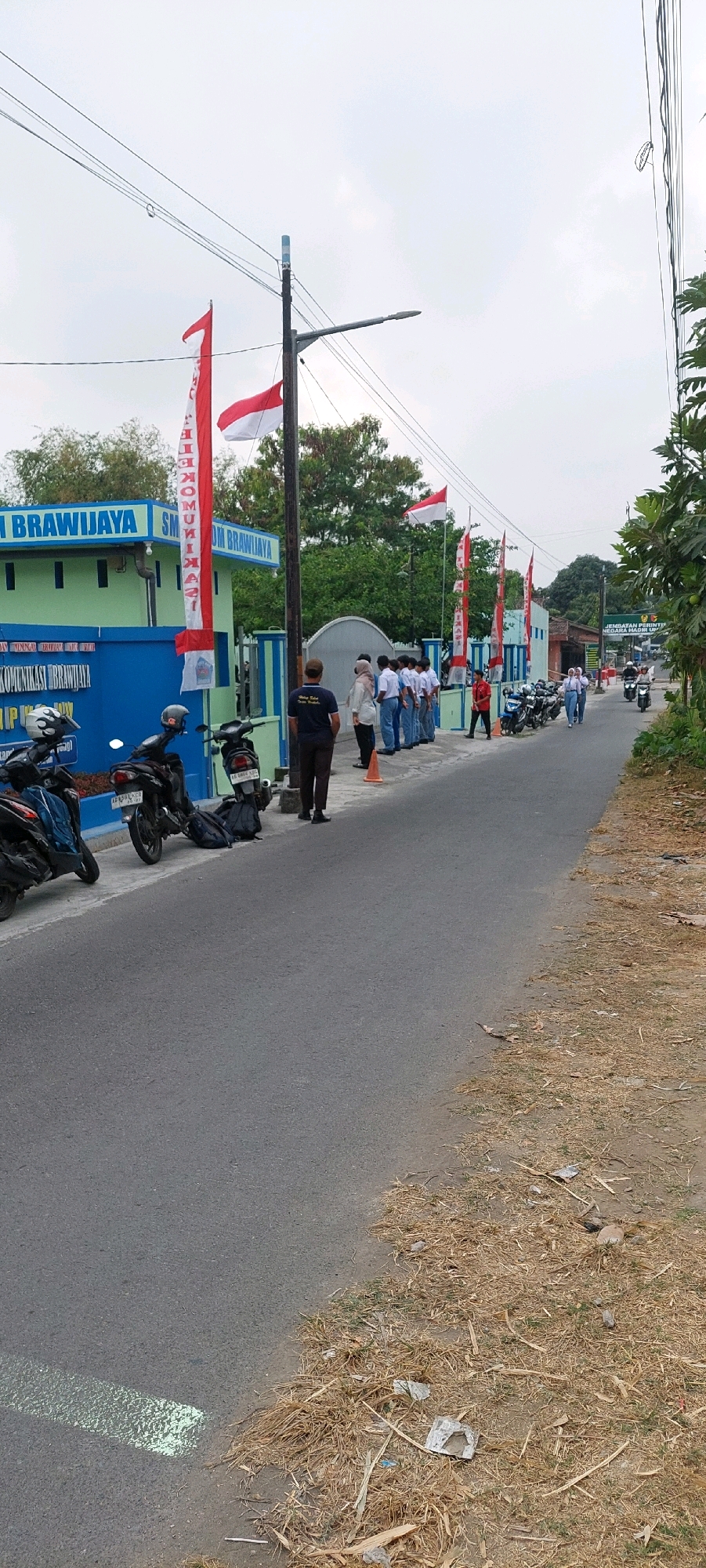 In an outdoor scene, a group of students, identifiable by their white shirts and dark pants or skirts, are standing in a line facing a green and blue building. The building has "SM BRAWIJAYA" written on it in large blue letters, and appears to be a school. Several red and white Indonesian flags are visible, some on poles and some fluttering on a building. Motorcycles are parked along the side of the road in front of the building, and a few are also seen on the road further down. The road is paved and an asphalt surface. Dry, light-colored grass and debris are present along the side of the road. The sky is overcast.