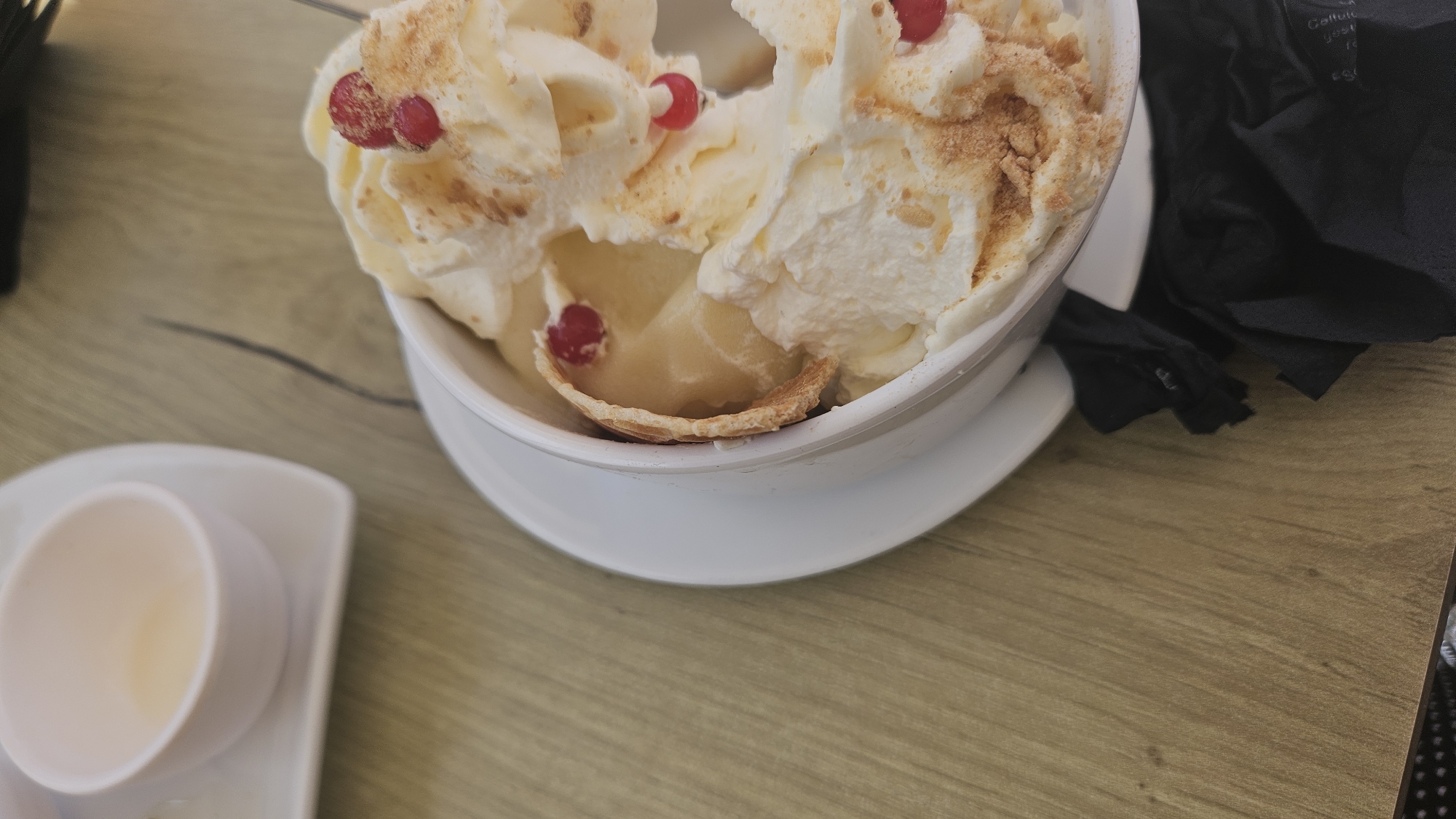 A close-up, high-angle shot shows a white bowl filled with a dessert, possibly ice cream or a parfait, topped with whipped cream and garnished with red berries and a wafer. The bowl rests on a white saucer, which is placed on a wooden table. To the left of the bowl is a small, white dish containing a light-colored liquid, possibly a sauce or drink, also on a white saucer. A black napkin is visible to the right of the bowl. The background is a wooden table surface with visible grain. The scene is indoors, likely in a cafe or restaurant in Saint-Sauveur-de-Montagut, France, given the context provided, although the image itself does not contain explicit location markers. There are no people visible in the frame. The lighting appears to be natural or ambient, casting soft shadows.