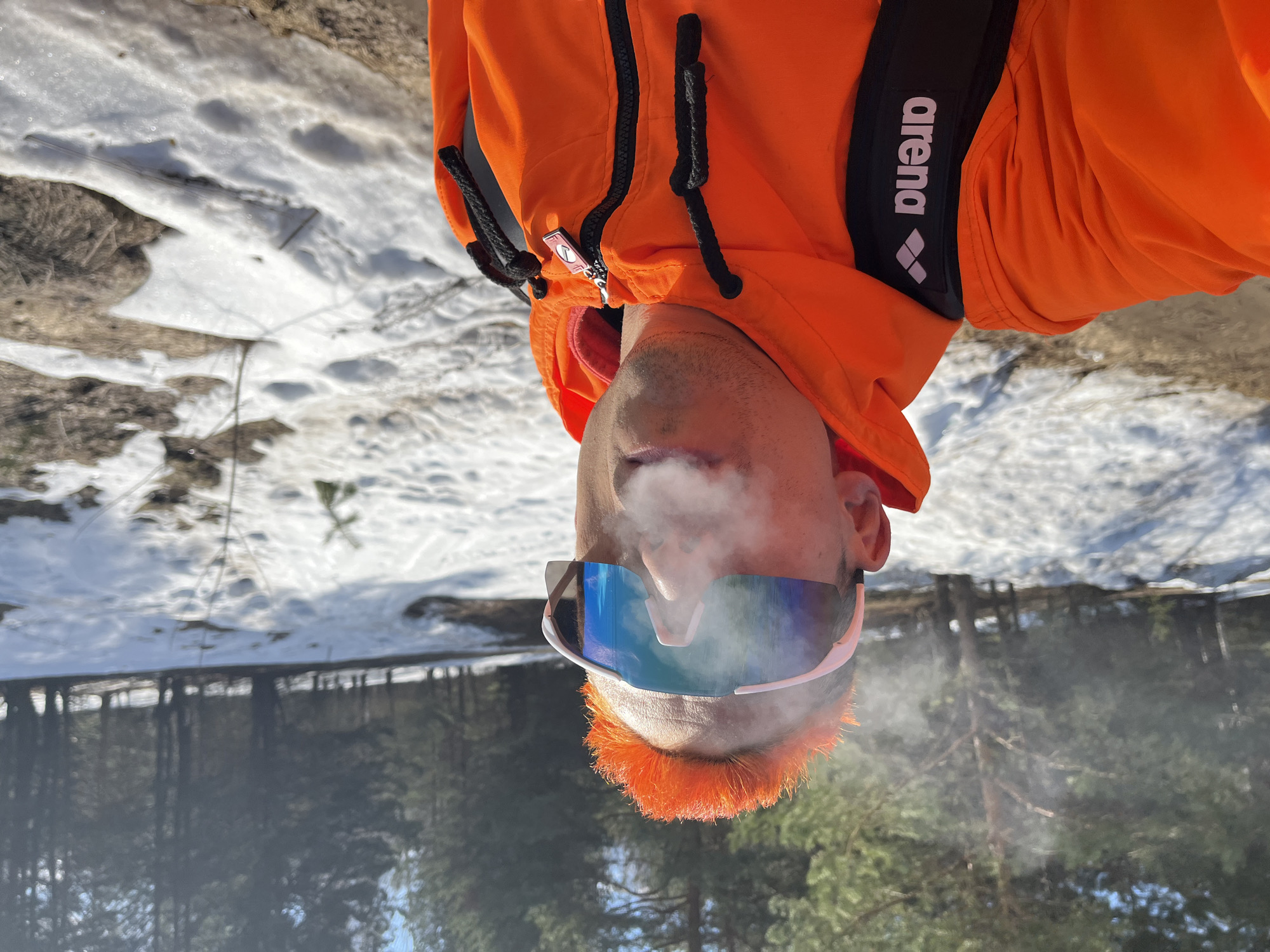 The image shows a man in Chaikovsky, Russia, standing outdoors in a snowy area, likely in a forest or park. He is wearing an orange jacket with a black "arena" strap and white sunglasses. He has orange hair and appears to be exhaling vapor, possibly from a vaping device. The background includes trees, snow patches, and possibly a body of water reflecting the trees. The image is oriented upside down.
