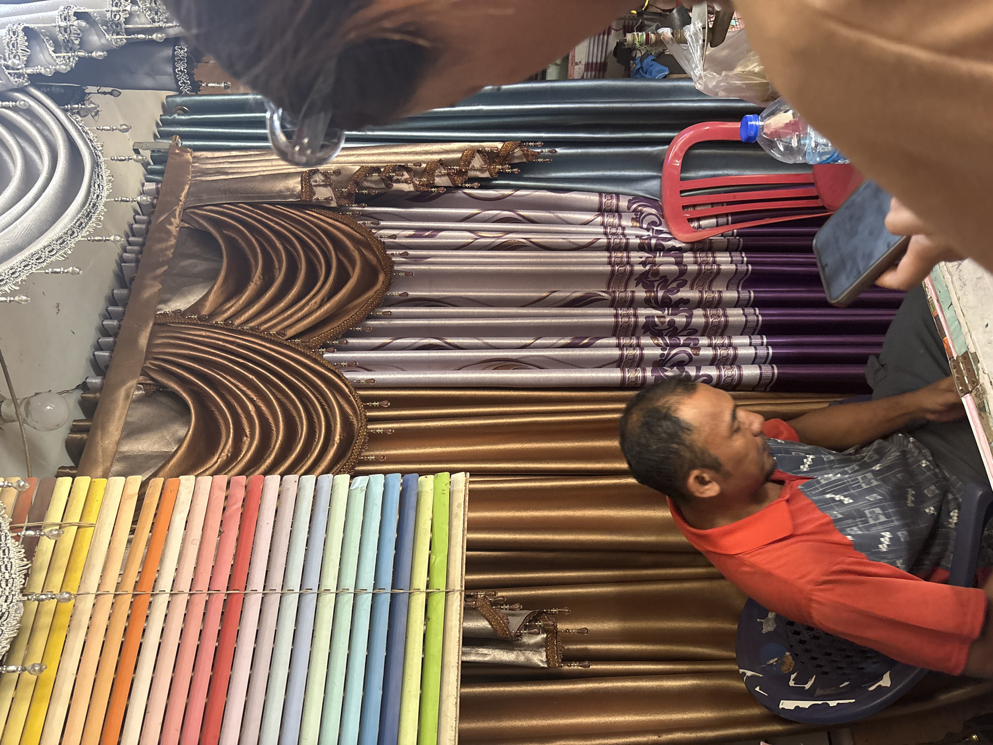A man sits in a blue plastic chair in what appears to be a shop selling curtains and blinds. He is wearing an orange polo shirt with a gray and white patterned insert on the chest and right side. The man is looking down, possibly at his phone, which is not clearly visible. Behind him, various curtains and blinds are displayed, featuring a variety of colors and textures, including a row of multi-colored blinds on the left and ornate gold and purple curtains in the background. A red plastic chair is visible to the right of the man. A plastic water bottle and a mobile phone are on a table in front of him. The scene suggests a retail environment, likely a fabric or home decor store in North Jakarta, Indonesia.