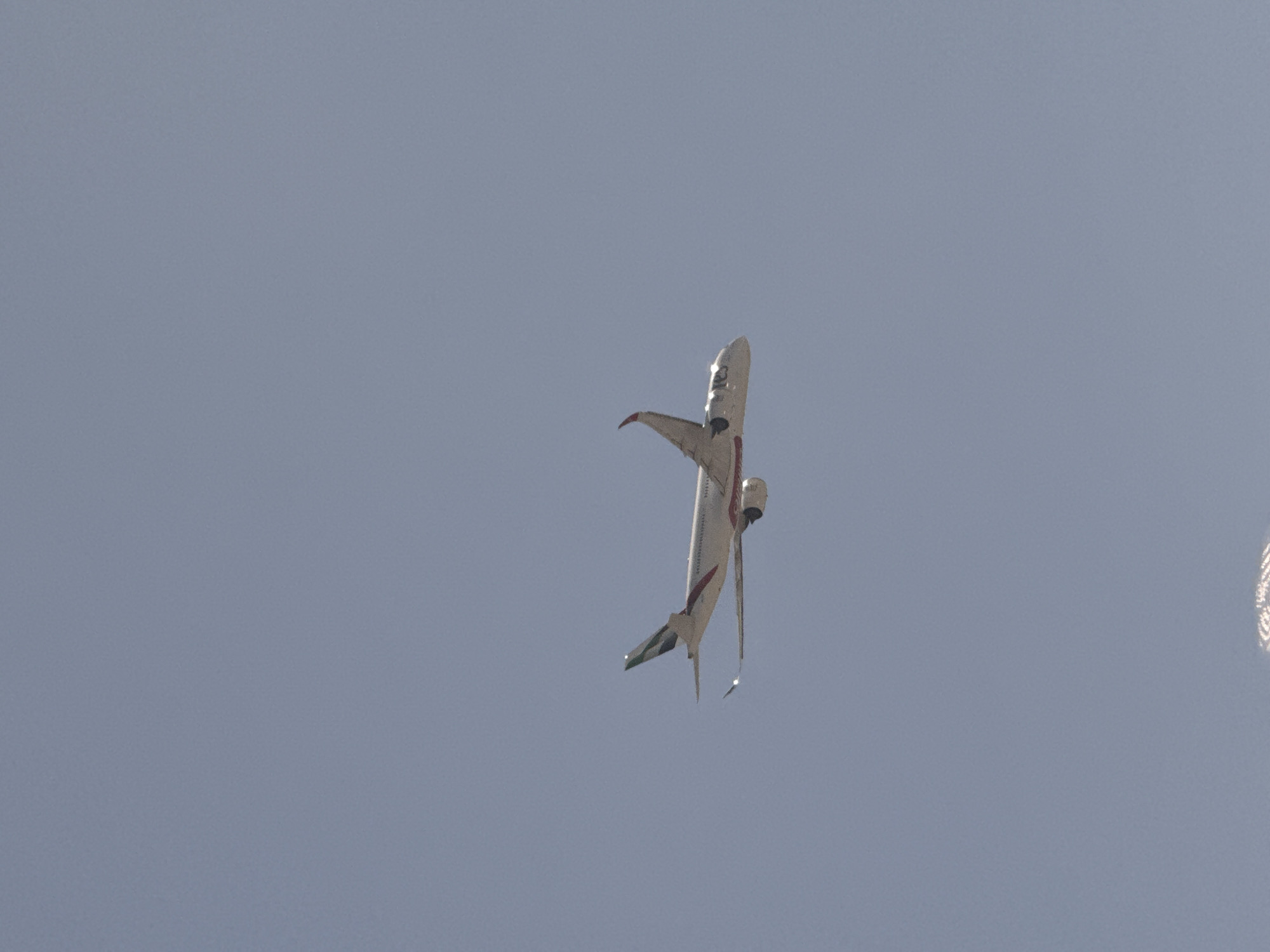 An airplane is flying in a clear, pale blue sky. The aircraft is white with red and green accents. Its wings are extended, and the engines are visible beneath them. The plane is angled slightly, with the nose pointing to the left and the tail to the right. The image appears to be taken from below, looking up at the aircraft. The lower portion of the image shows a distorted, blurry reflection that appears to be of a building or structure, possibly indicating the presence of water or a reflective surface below. The sky suggests it is daytime. There are no people or other objects clearly identifiable in the scene.