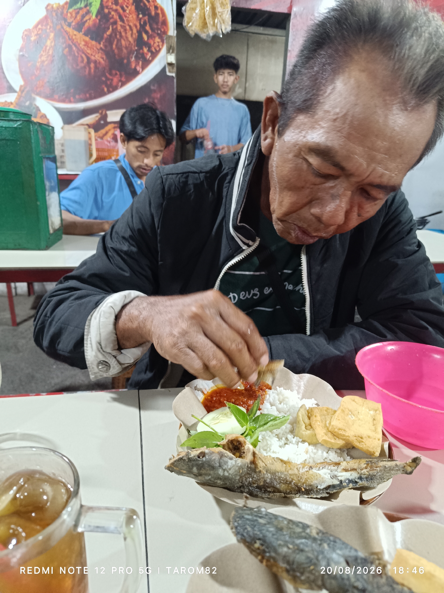 An aging East Asian man, with sweat glistening on his brow, is eating a meal at an outdoor food stall in Bringinbendo, Indonesia. He is wearing a black jacket over a dark t-shirt. In front of him is a plate of white rice, a piece of fried fish, some fried tofu, sliced cucumber, and a dollop of red chili sauce. He is using his right hand to pick up food from the plate. A glass of iced tea sits to the left of the plate. In the background, two younger East Asian men are visible. One is wearing a blue t-shirt and is looking down at something. The other is further back, wearing a light blue t-shirt. The food stall appears to have a mural of a dish on the wall behind the man in the blue t-shirt. The timestamp on the image reads "20/08/2026 18:46".