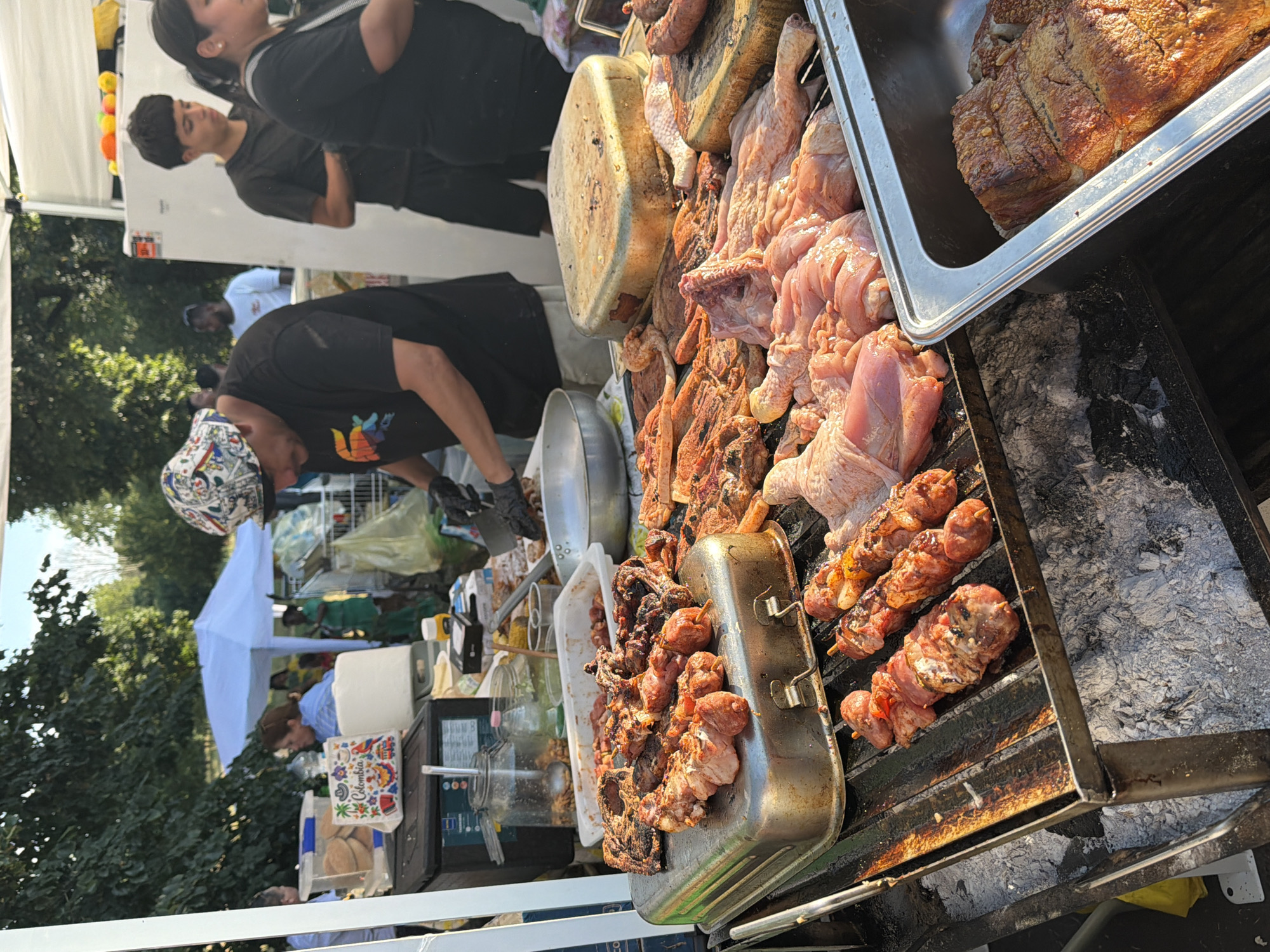 The image depicts an outdoor food stall, likely at a public event or market. In the foreground, a variety of raw and cooked meats are displayed on a grill. Skewers of meat, along with larger cuts of meat, are visible. The grill appears to be fueled by charcoal. Behind the grill, a person wearing a black t-shirt and a patterned hat is seen preparing food, wearing black gloves and holding a knife. Other individuals are present in the background, including a young man in a black t-shirt and a woman in a black t-shirt. To the left of the scene, there are various containers, bottles, and a cooler. In the far background, a white tent and trees are visible, suggesting an outdoor setting. The image does not contain any specific landmarks or details that would definitively place it in the city of Segrate, Italy.