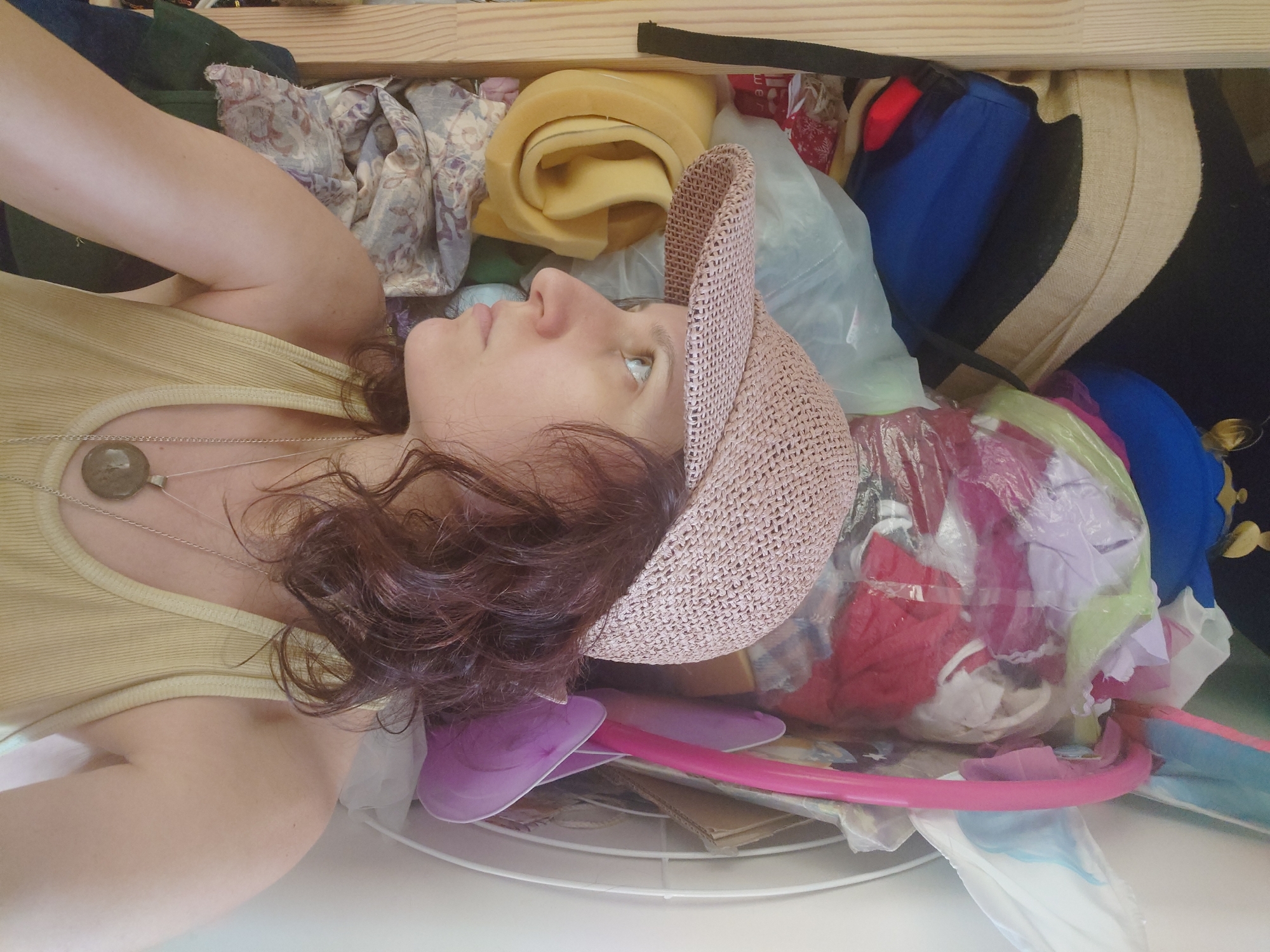 In the city of Kommunarka, Russia, a woman is positioned in the foreground, turned to her left, looking off into the distance. She is wearing a light yellow tank top and a light pink woven baseball cap. A silver chain with a round, dark pendant rests on her chest. Her dark, curly hair is visible beneath the cap. The background is cluttered with various items, including rolled-up yellow foam, folded fabric with a floral pattern, blue and black bags, and a pile of colorful items contained within plastic bags. A pink plastic hoop is also visible. The environment appears to be an indoor space, possibly a storage area or a cluttered room.