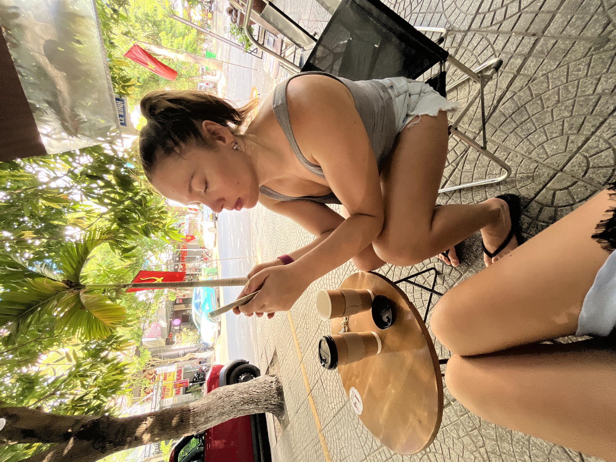 An outdoor, street-level scene in Đà Nẵng, Vietnam, features two individuals at a sidewalk cafe. In the foreground, the legs of the photographer are visible, clad in light-colored shorts with a black lace trim. A small, round, light-wood-topped cafe table with a black metal base occupies the mid-left. On the table are two cardboard-sleeved paper cups, one with a black lid and one open, alongside a small black saucer.
Seated opposite the photographer on a black mesh folding chair with a metal frame is a female with light skin, wearing a grey tank top and light blue ripped denim shorts. Her brown hair is pulled back into a high ponytail. She is looking downwards with her eyes closed or partially shut, intently holding a gold-colored smartphone with both hands. A purple wristband is visible on her right wrist, and black flip-flops are on her feet.
The background reveals a bustling street scene under dappled sunlight filtering through lush green foliage from several trees, including a prominent tree trunk in the left foreground and palm trees. A red Ford Bronco-like SUV is parked roadside on the left. Across the street, vehicles including a light blue sedan, a white sedan, and motorcycles are visible. The far side of the street features commercial buildings with multiple signs, notably several displaying "MASSAGE" and "VIP" in red and white lettering. A blue sign with white text, partially reading "LÊ BÌNH," is also visible. Two red Vietnamese flags are displayed; one is the national flag with a yellow star, and the other, on a pole next to a palm tree, features a yellow hammer and sickle, indicative of the Communist Party. The sidewalk beneath the cafe area is paved with grey and tan interlocking bricks in a curved pattern.