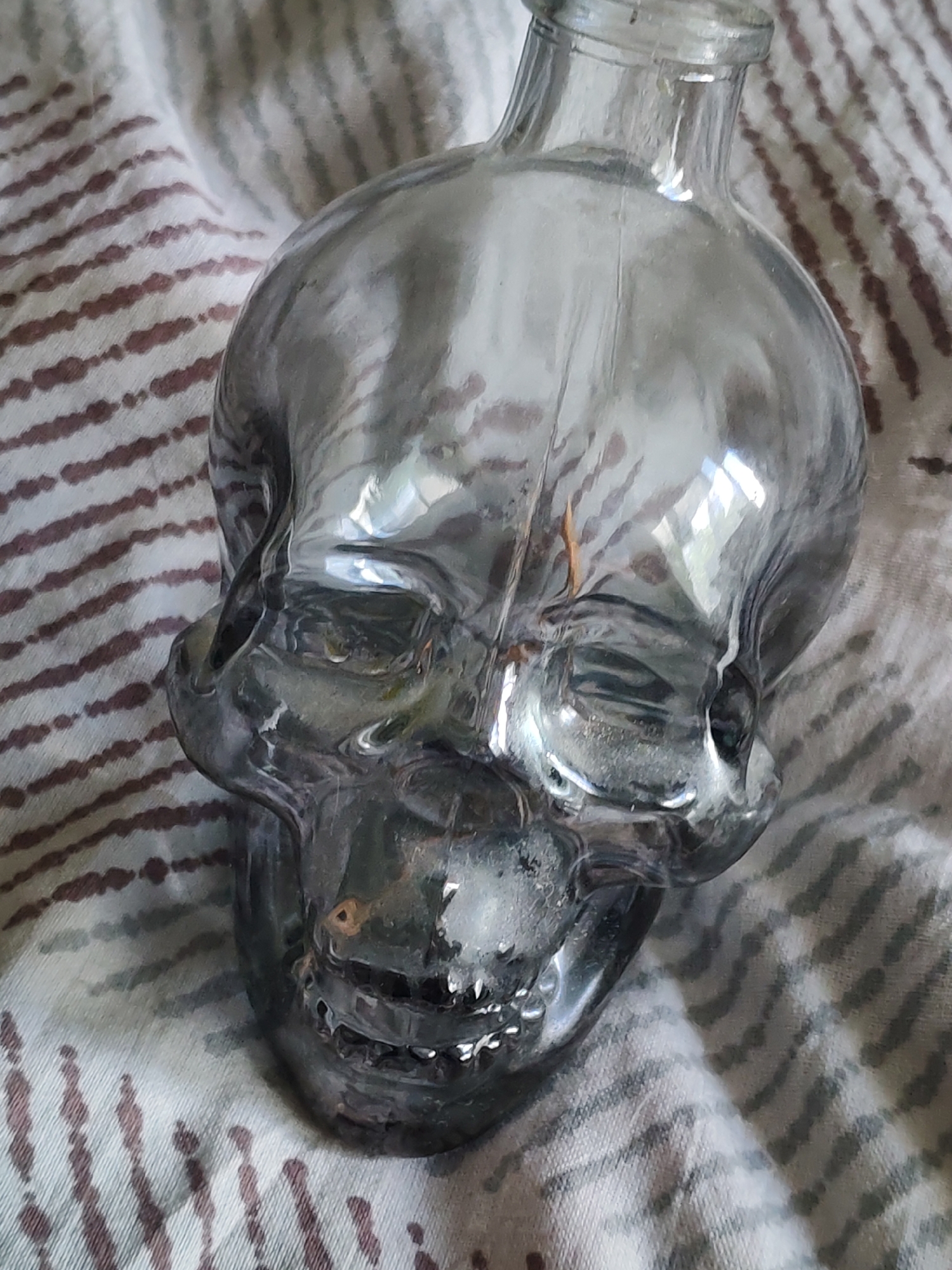 A close-up shot of a clear, skull-shaped glass bottle on a patterned fabric. The bottle is empty and resting on the fabric, which has horizontal stripes in varying shades of grey and brown. The image does not provide enough detail to discern the location or any information about people or actions.